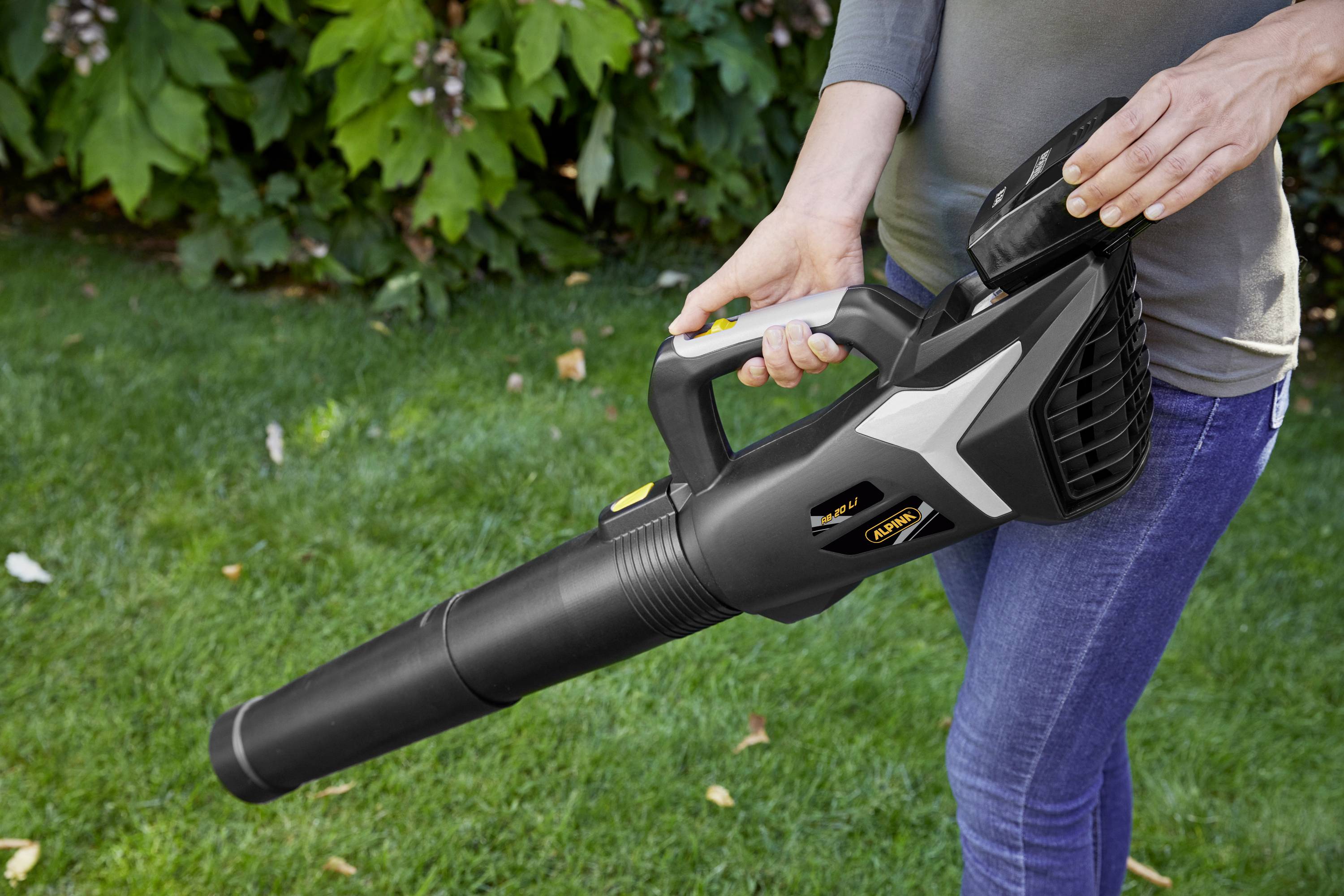 A person is holding a black cordless leaf blower in the garden. The blower is being operated with both hands. Green plants are visible in the background.
