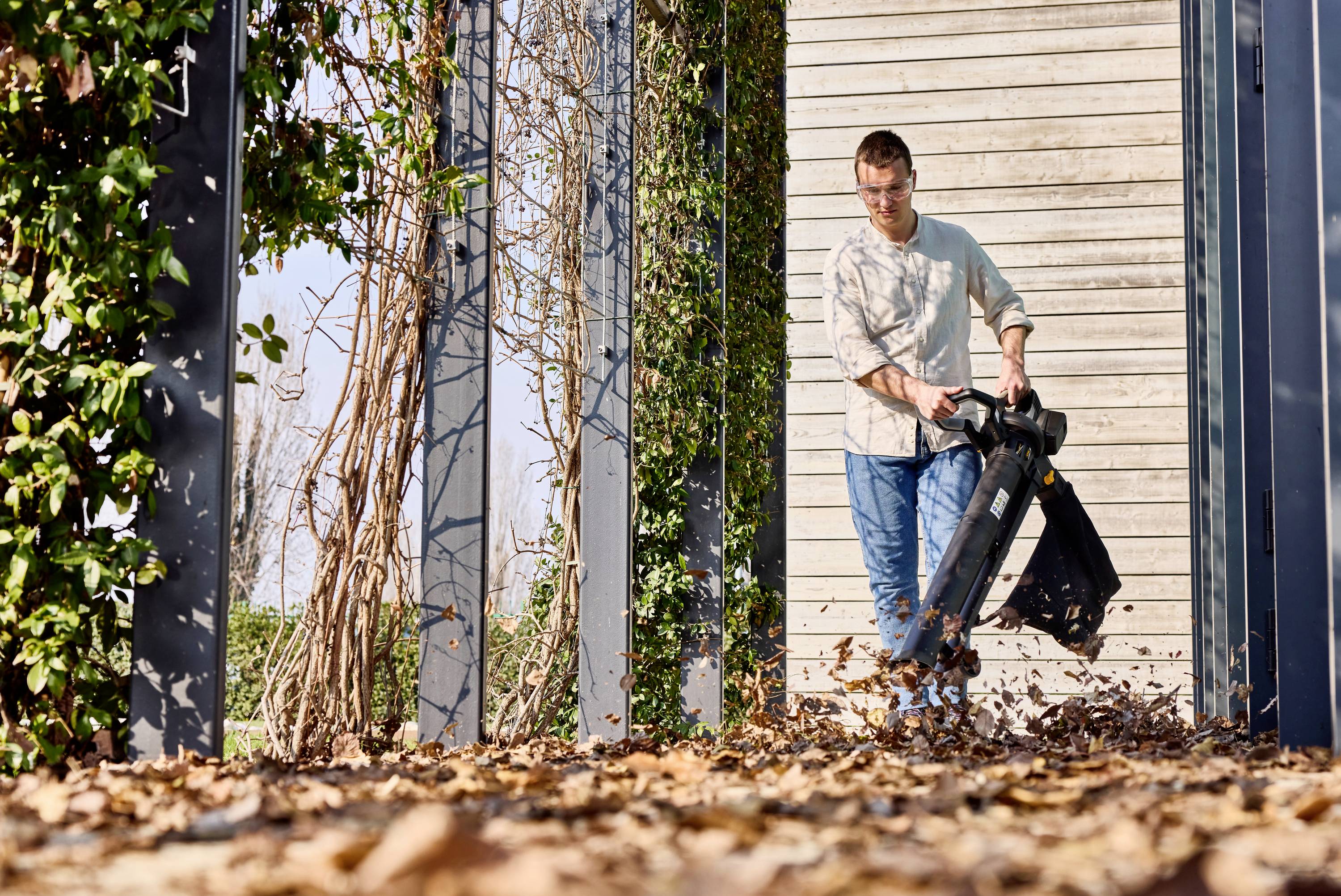 A man is gathering dry leaves with a leaf blower in a yard. Columns and ivy are visible in the background.
