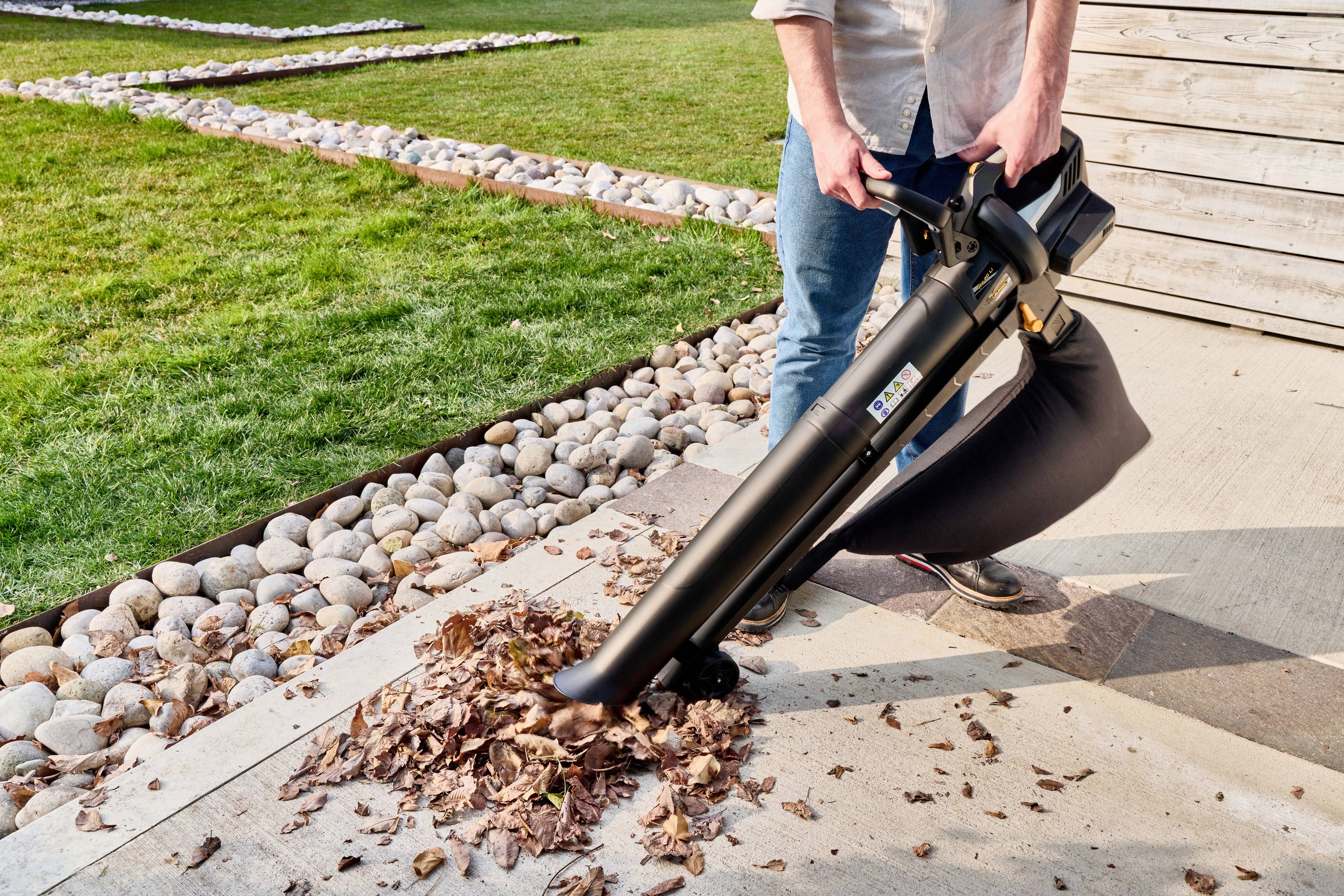 A person is using a leaf blower to remove leaves from a paved surface. In the background, there is grass and stones.