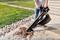 A person is using a leaf blower to remove leaves from a paved surface. In the background, there is grass and stones.