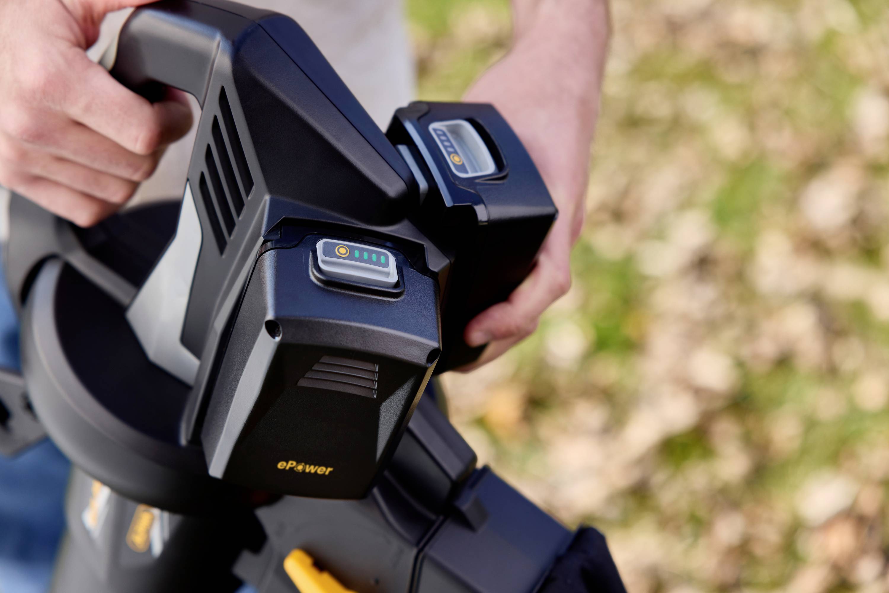 A person is holding a leaf blower and inserting a battery; background with autumnal leaves on grass.