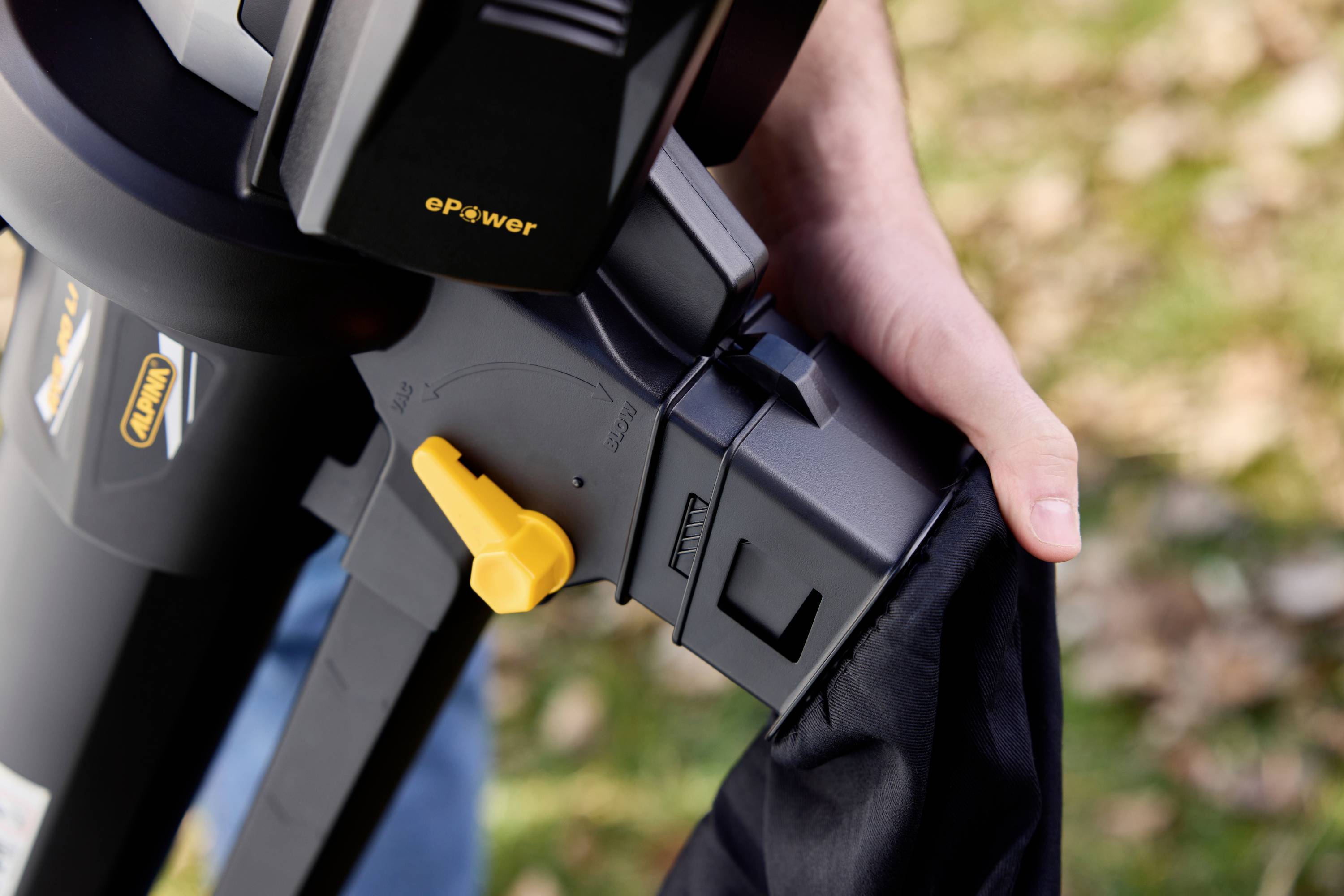 A person is holding a pipe with a yellow lever; it looks like part of a tool or device. Blurred leaves are visible in the background.