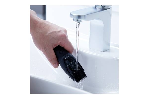 A hand holds an electric razor under running water from a tap in the sink.