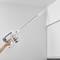 A person is using a cordless vacuum cleaner with a long wand to clean a corner near the ceiling.
