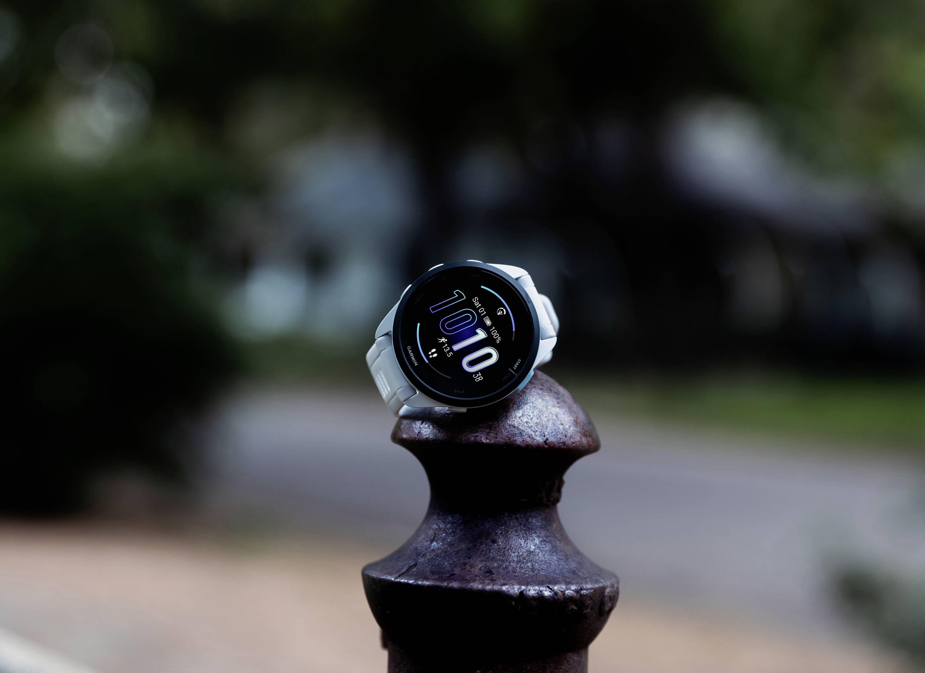 A smartwatch with a white strap displays the time as 10:10 on a round stand outdoors. The background is blurred.