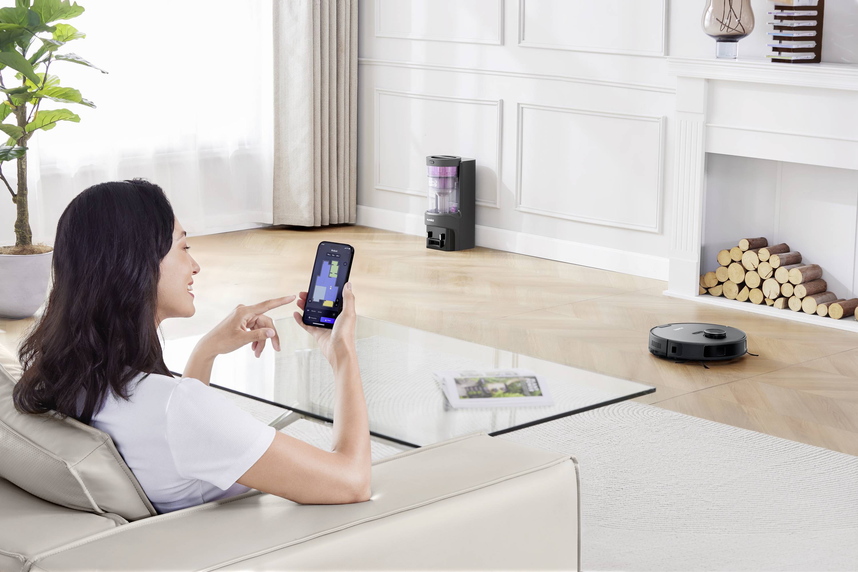 A woman controls a robotic vacuum cleaner via smartphone in a modern living room. Plants and a fireplace are in the background.