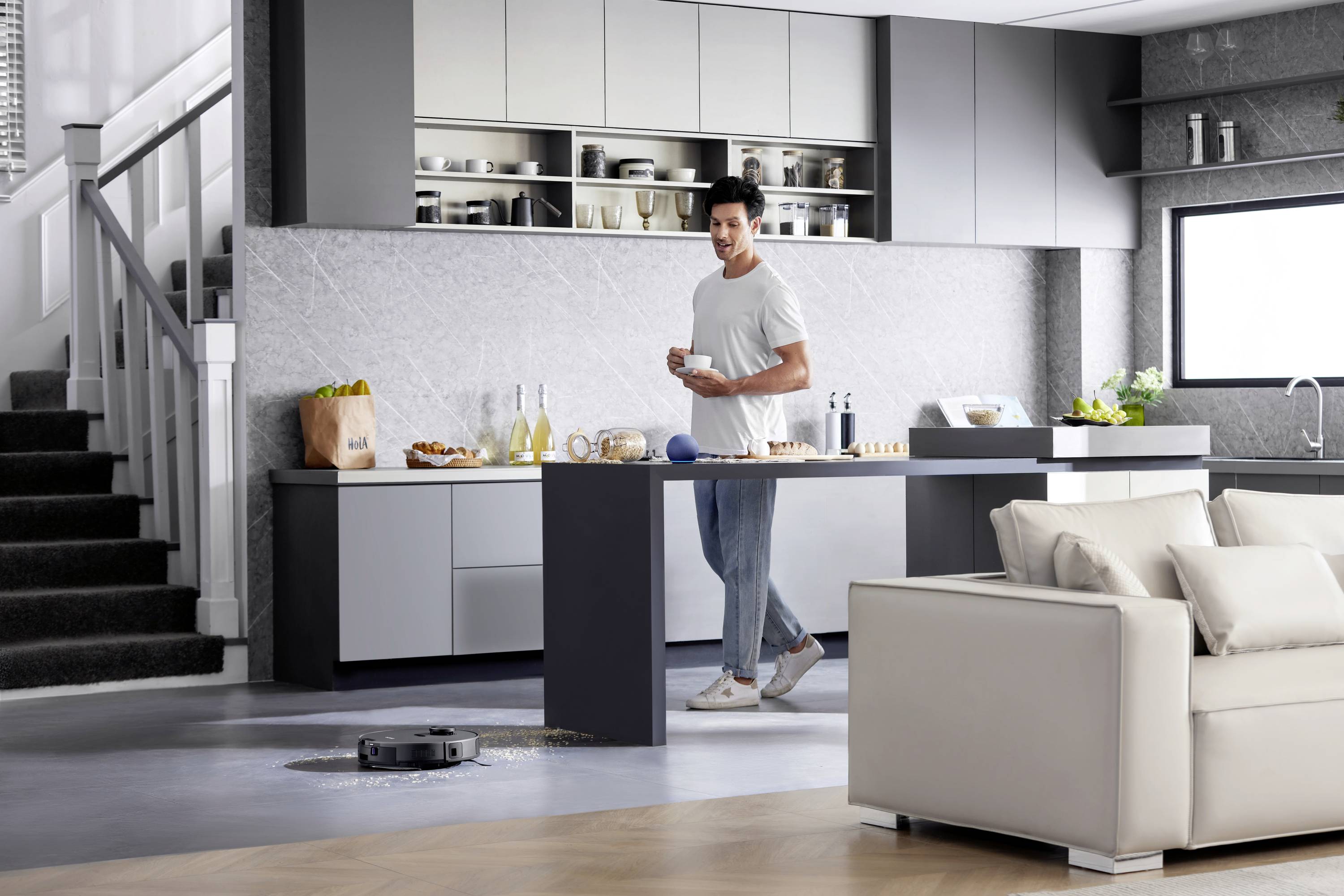 Man in a modern kitchen, standing next to a dining table. A robotic vacuum cleaner is cleaning the floor. Food preparation is in progress.