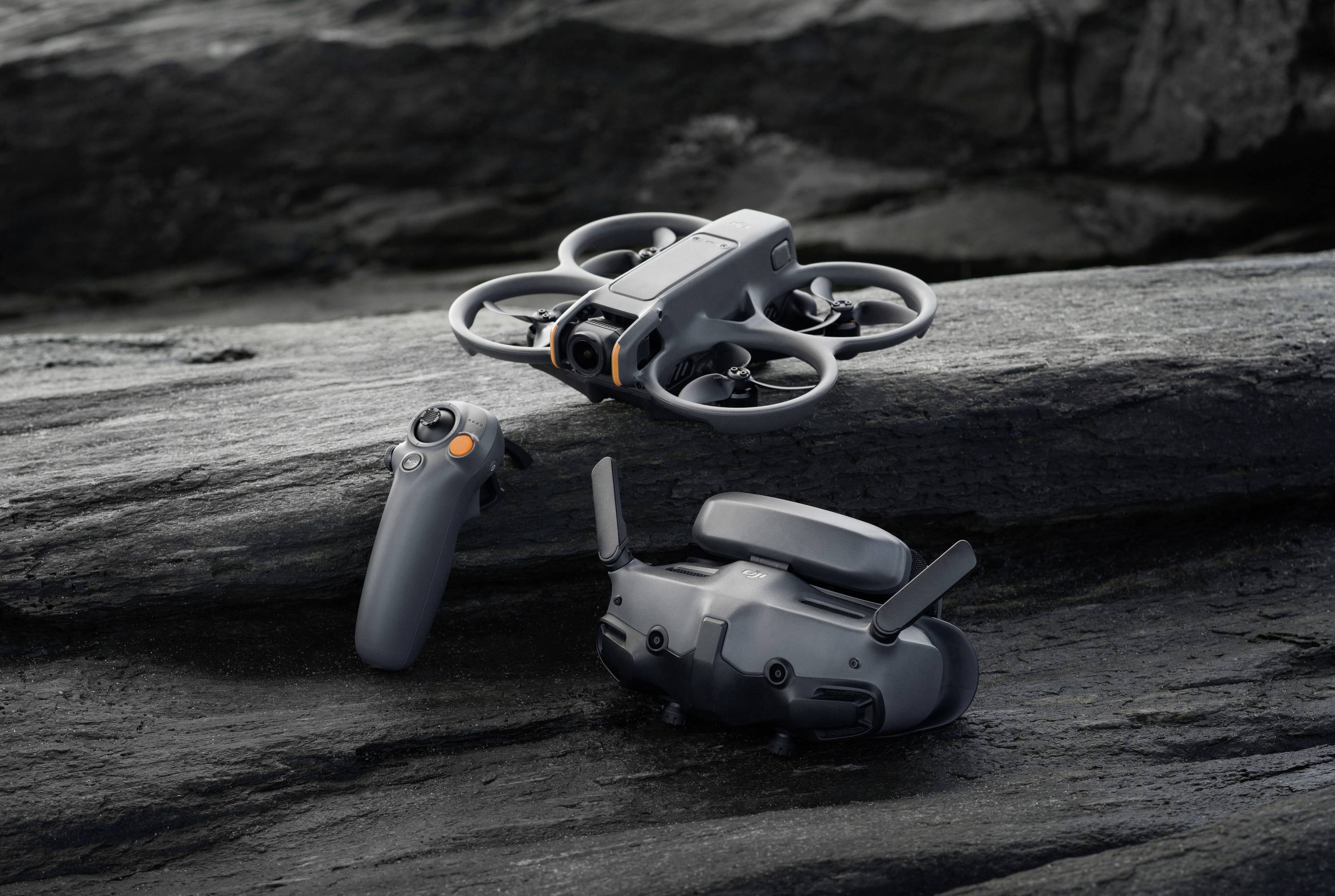 A grey drone hovers above a rocky surface. Beside it lie a remote control and a VR headset.