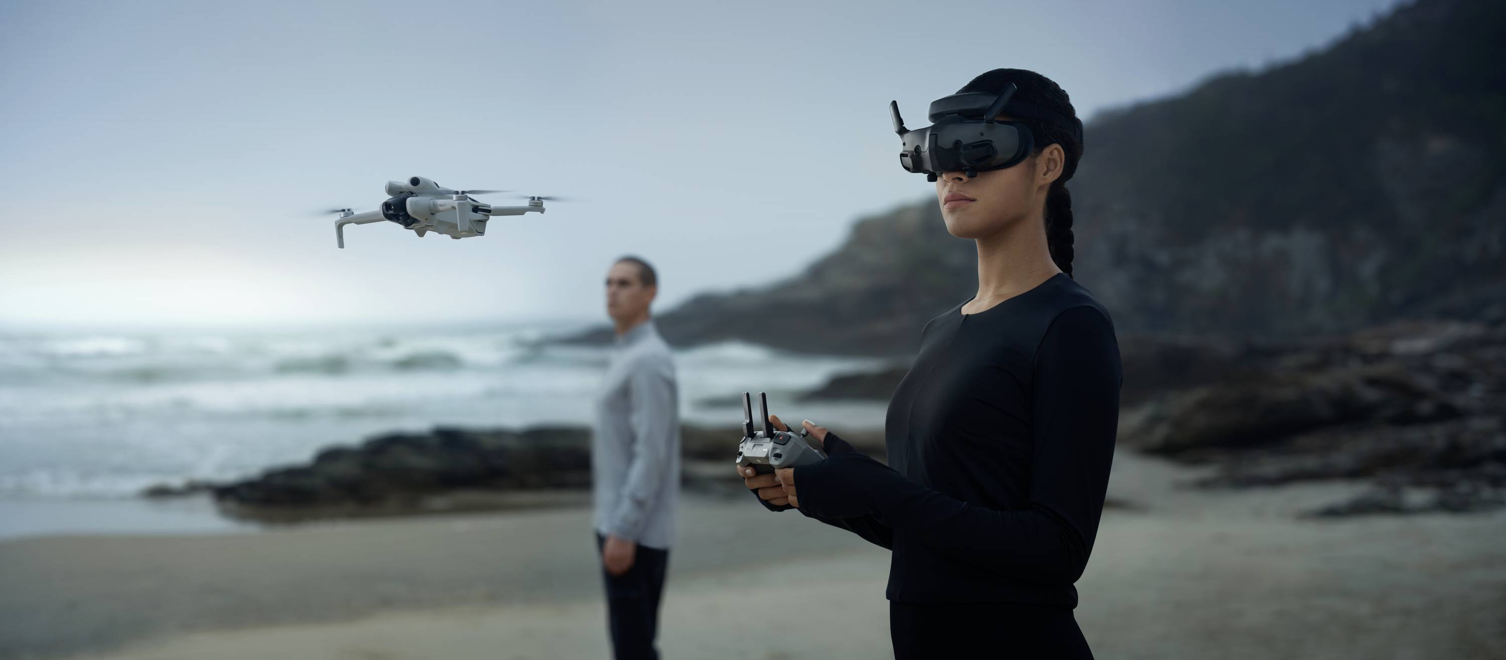A woman is controlling a drone with a virtual reality headset on a beach, while a man stands in the background.