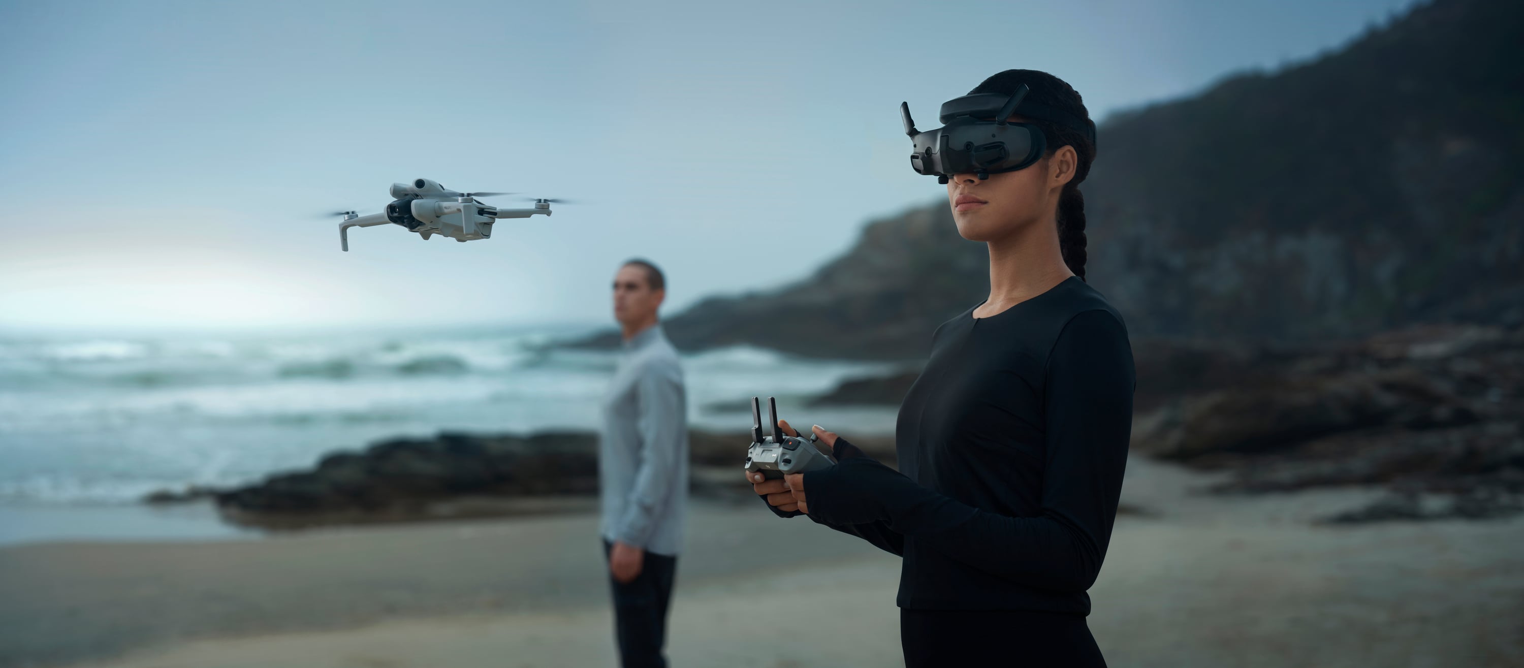 A woman is controlling a drone with a virtual reality headset on a beach, while a man stands in the background.