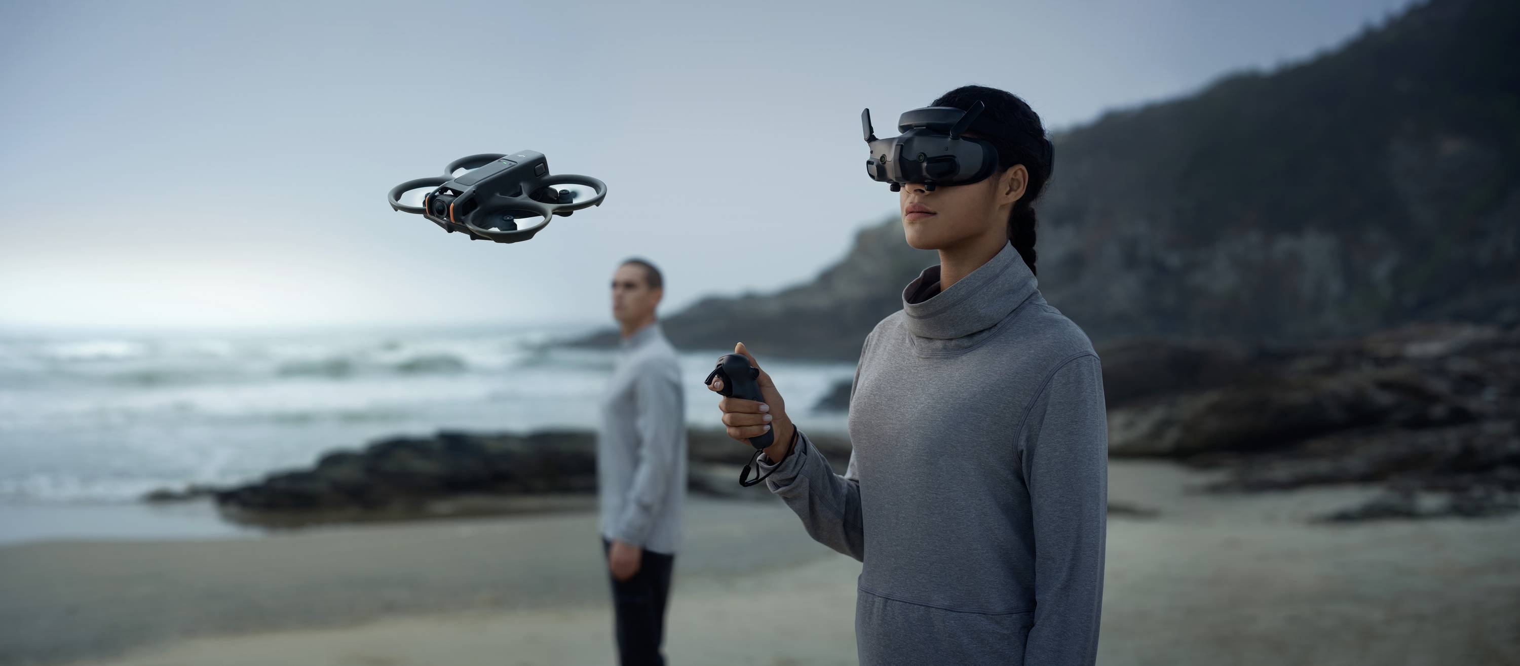A person is controlling a drone with a remote control and VR headset on the beach, with another person standing in the background.