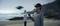 A person is controlling a drone with a remote control and VR headset on the beach, with another person standing in the background.