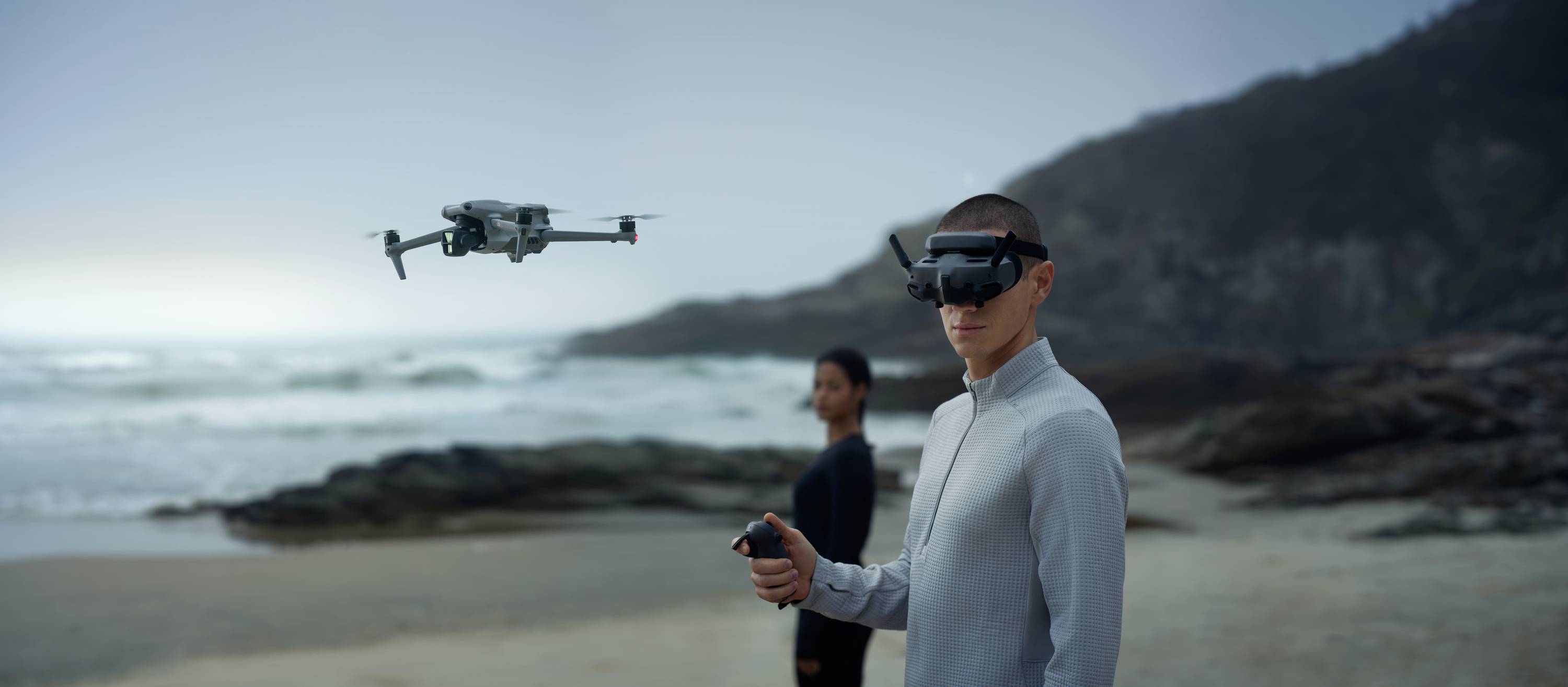 Two people wearing VR headsets are controlling a drone on the beach at sunset. The drone hovers above the sand with the sea in the background.