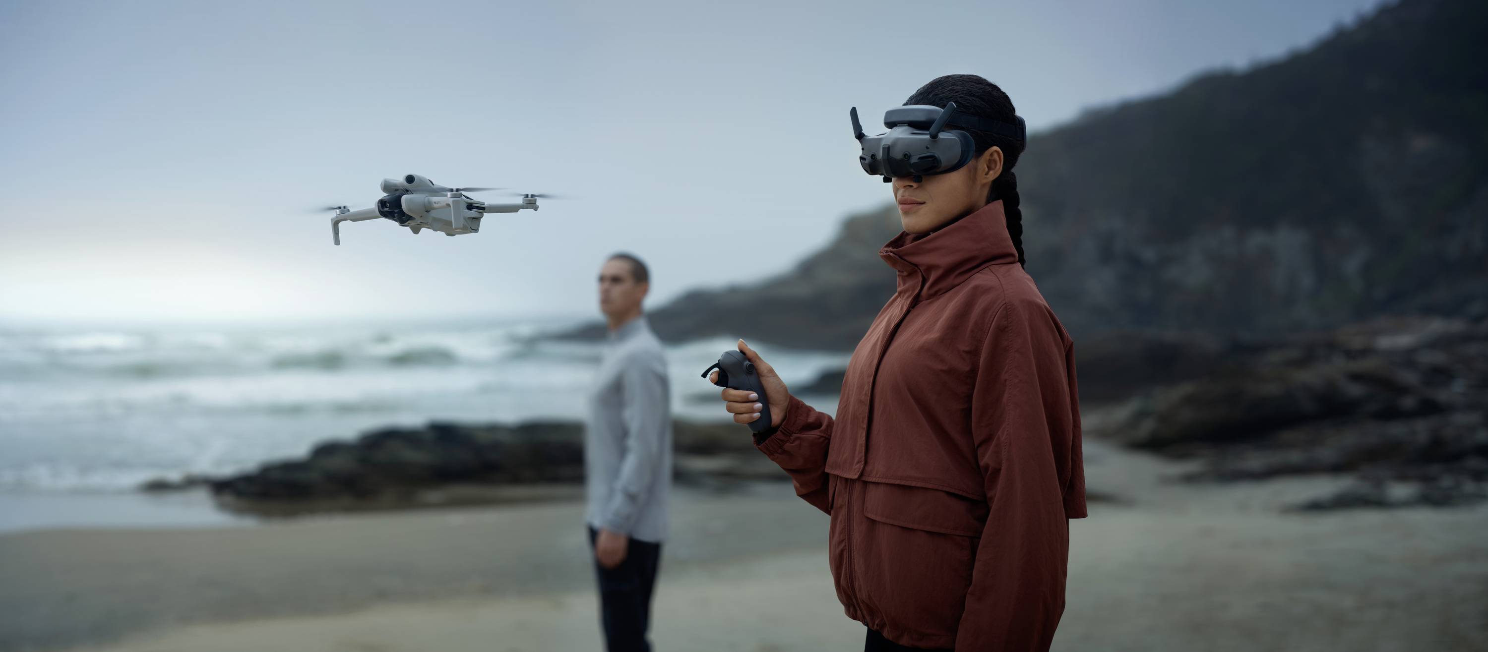 A person wearing a VR headset is controlling a drone on the beach. Another person stands in the background, looking out to sea.