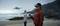 A person wearing a VR headset is controlling a drone on the beach. Another person stands in the background, looking out to sea.