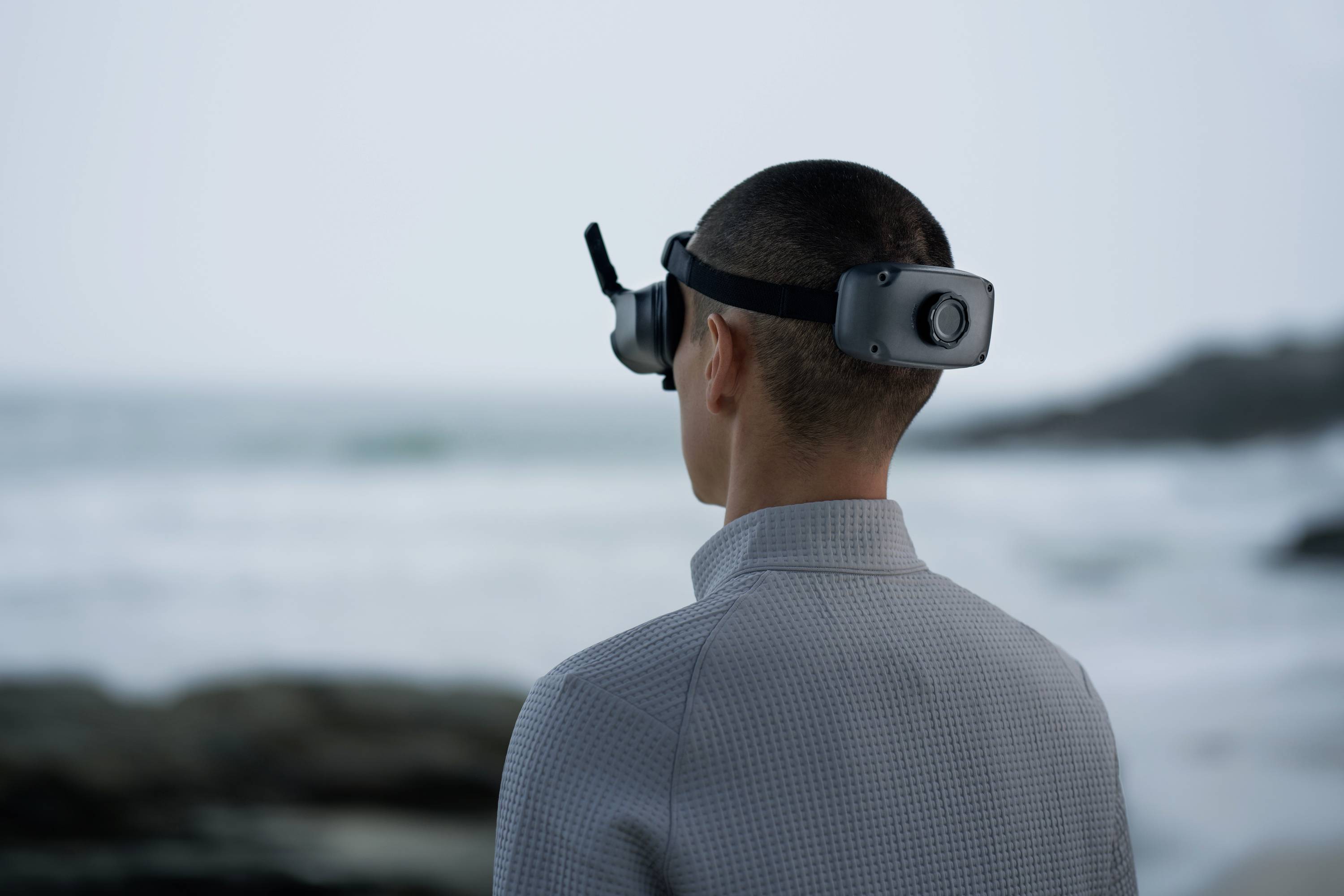A person gazes out to sea, wearing a VR headset. They stand on the beach, with waves crashing against rocks in the background.