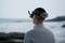 A person gazes out to sea, wearing a VR headset. They stand on the beach, with waves crashing against rocks in the background.
