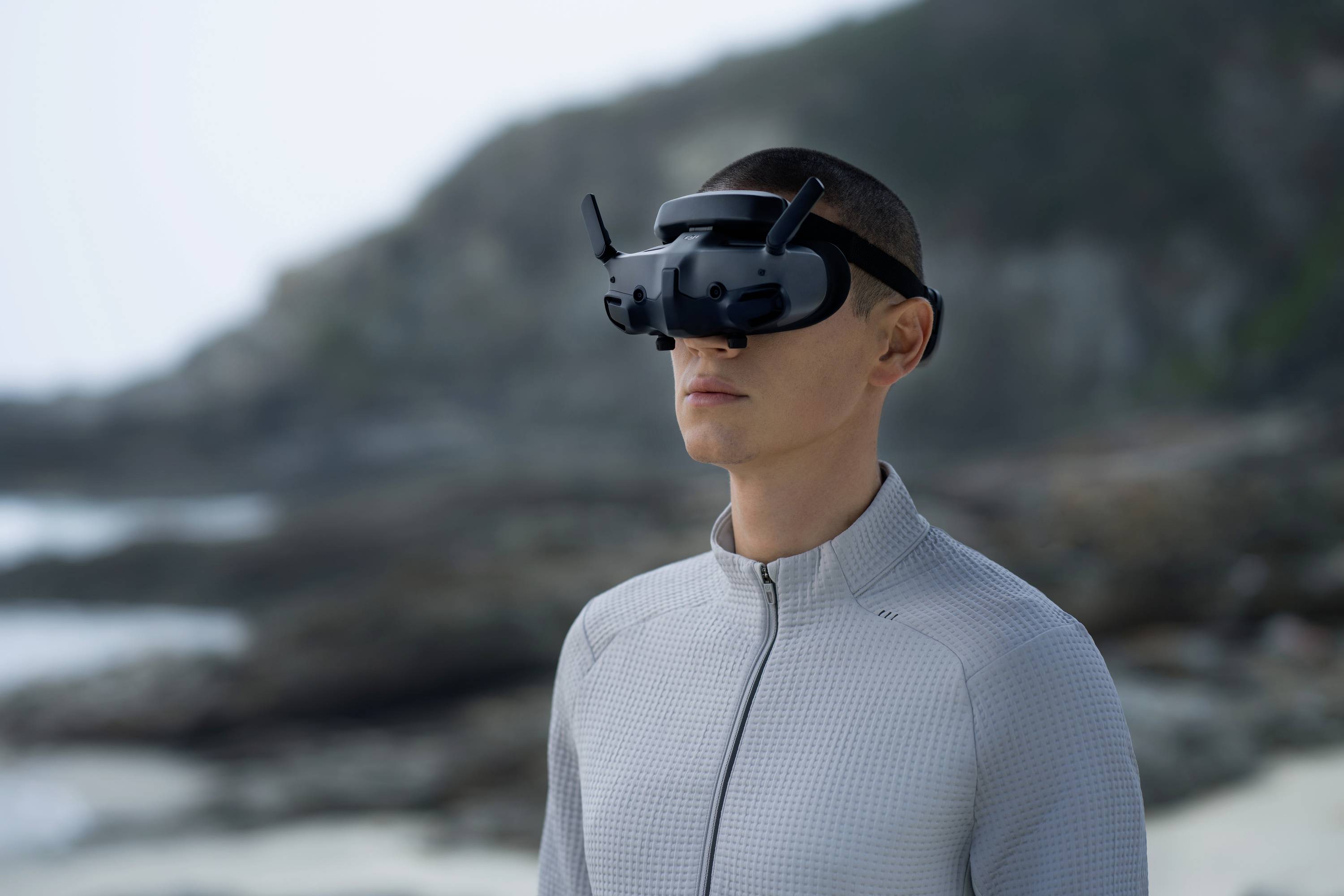 A person wearing a VR headset stands on the beach, looking straight ahead. Rocks and the sea are visible in the background.