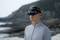 A person wearing a VR headset stands on the beach, looking straight ahead. Rocks and the sea are visible in the background.