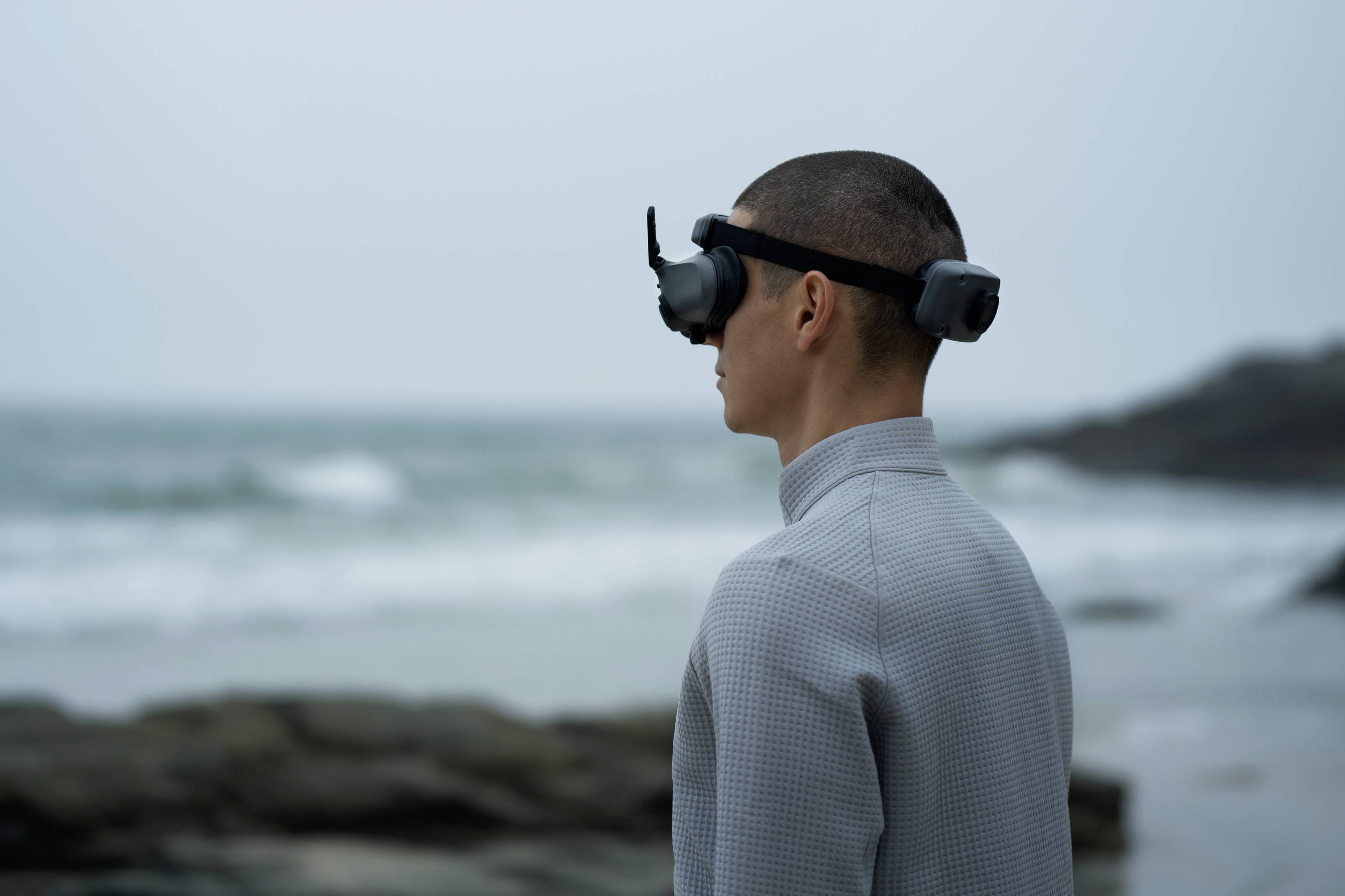 A person stands by the sea wearing a virtual reality headset. The background shows waves and a cloudy sky.