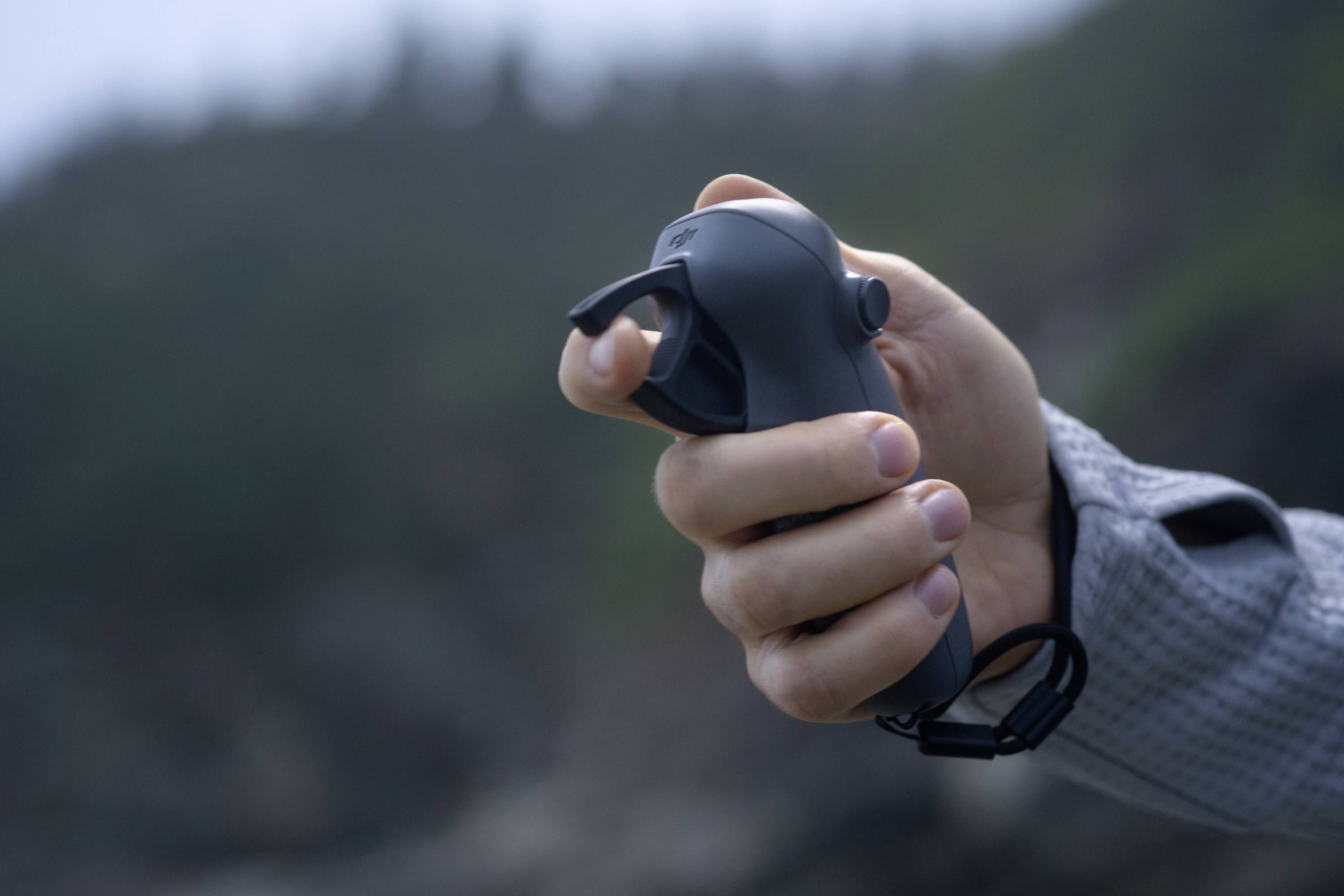 A hand is holding a grey handle with a trigger button, surrounded by a blurred, natural background.