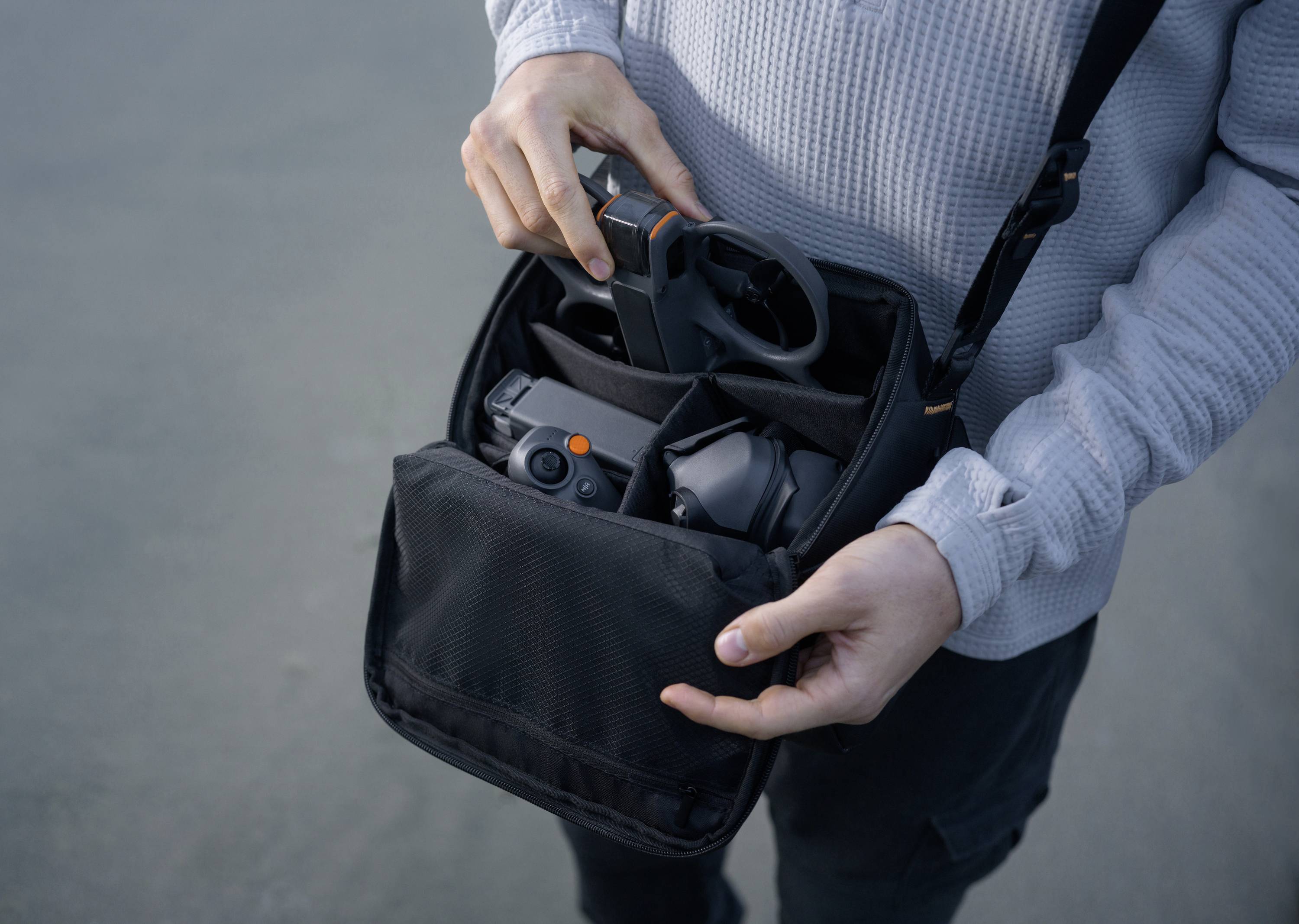 A person is holding an open bag with drone accessories, including a camera and a controller, in their hands.