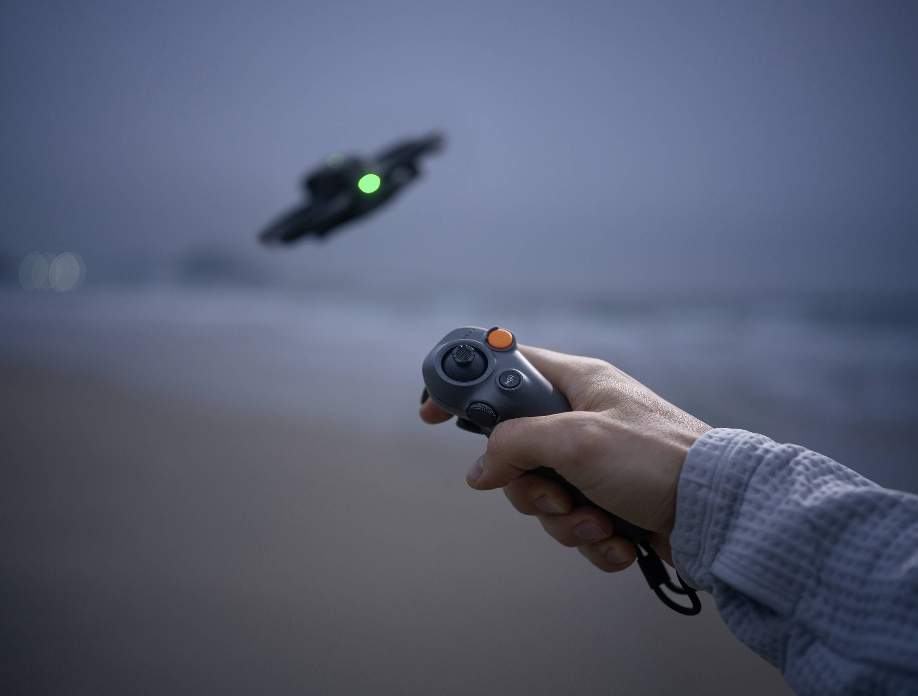 A hand controls a drone with a controller on the beach at dusk. The drone has a green light and is flying over the sea.