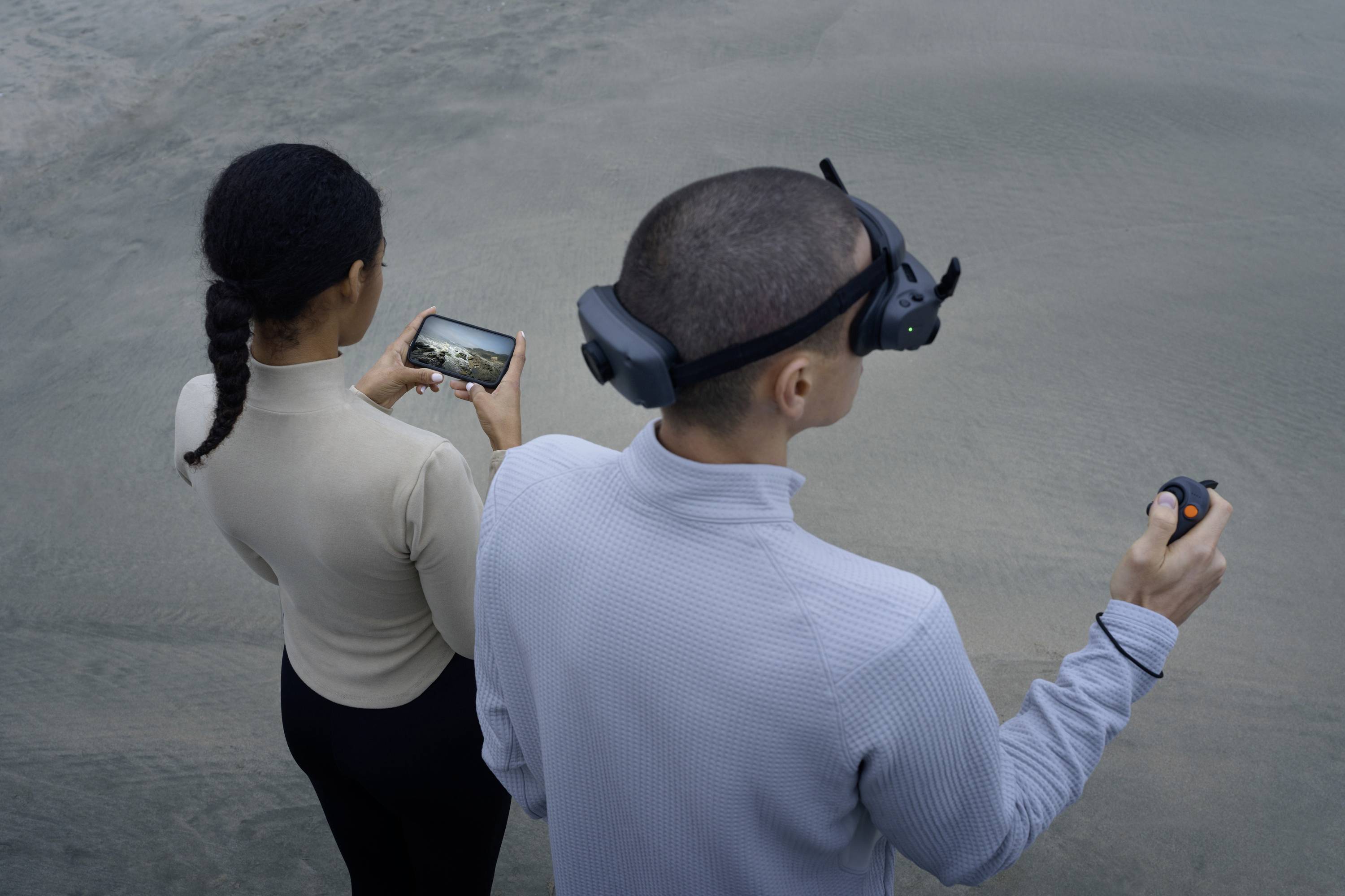 Two people are standing on sandy ground. A woman is holding a smartphone with a landscape display. A man is wearing a VR headset and holding a controller.