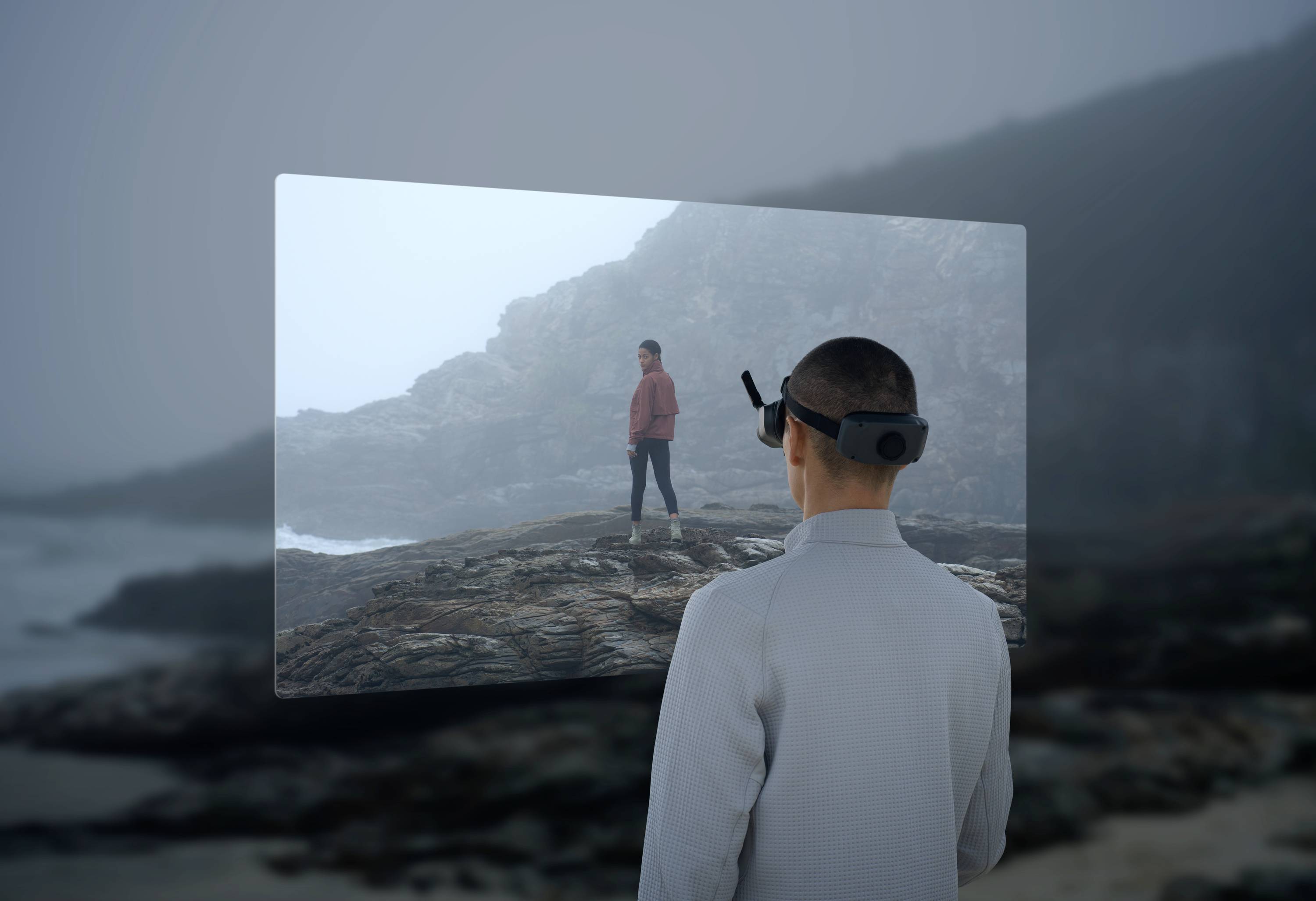 A person stands on the beach, gazing at a holographic projection of a human in a rocky coastal landscape.