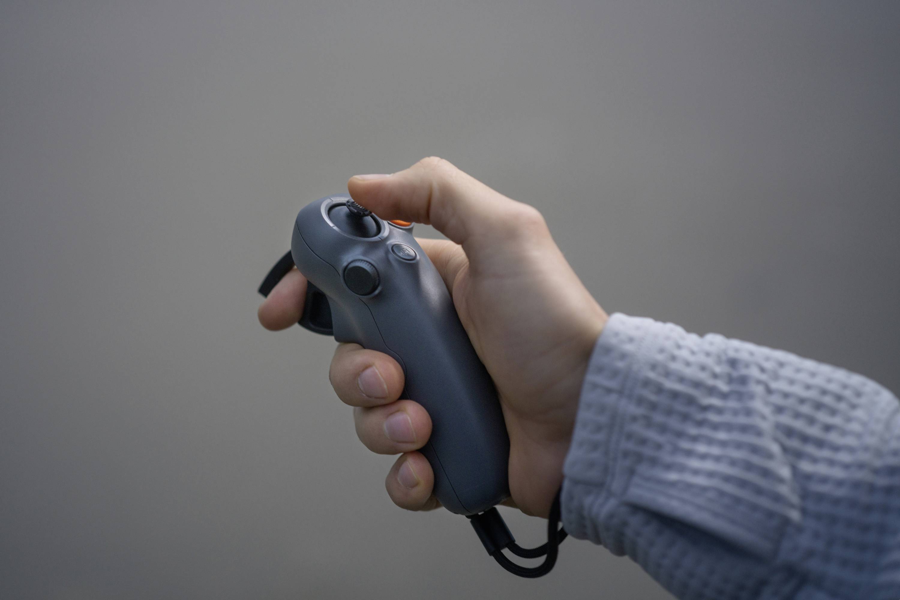 A hand is holding a grey controller with multiple buttons and a joystick. The background is blurred and neutrally coloured.