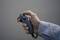 A hand is holding a grey controller with multiple buttons and a joystick. The background is blurred and neutrally coloured.