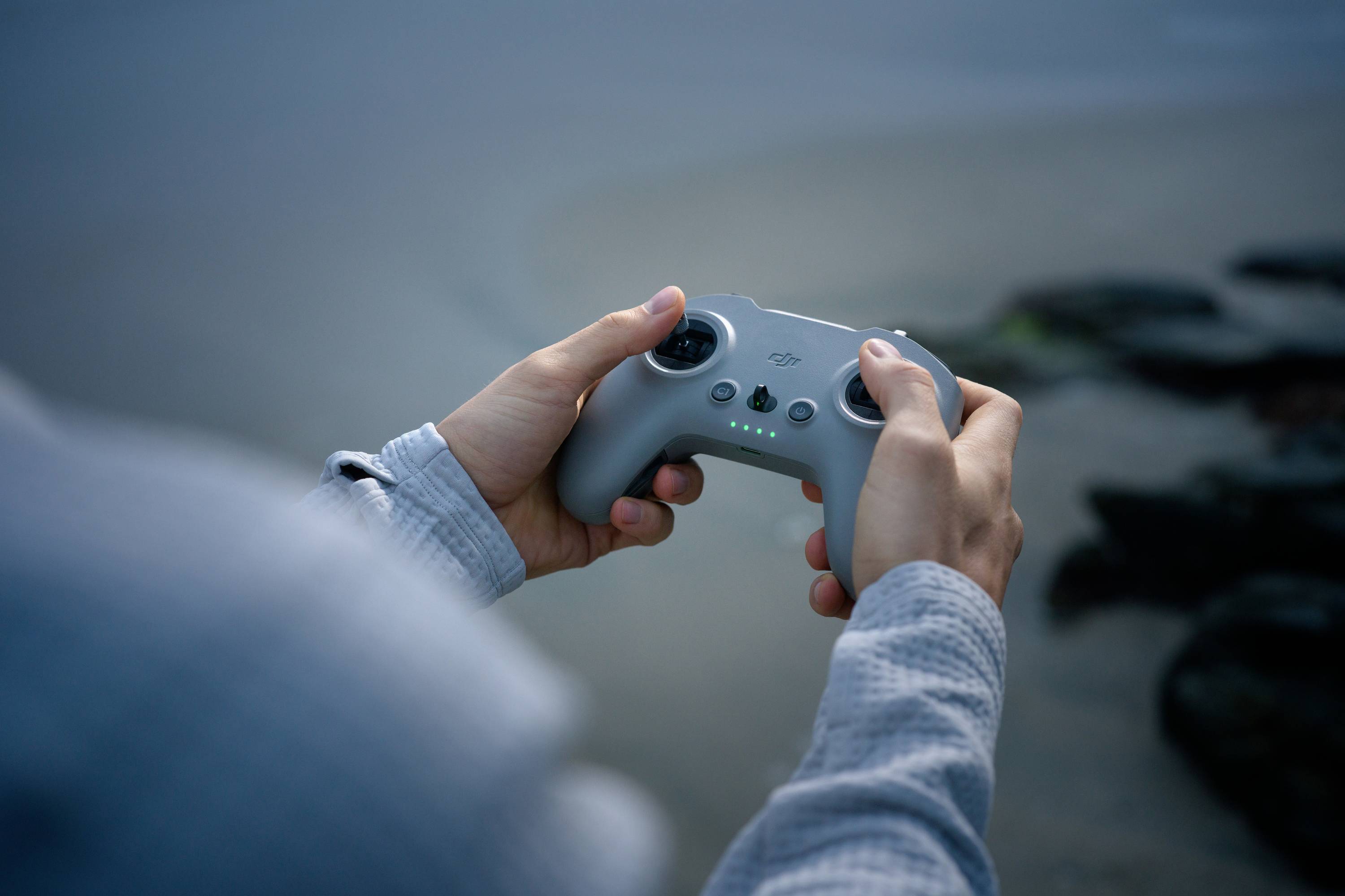 A person is holding a grey remote control outdoors with a blurred background. Controlling a device; focus on the hands and the controller.