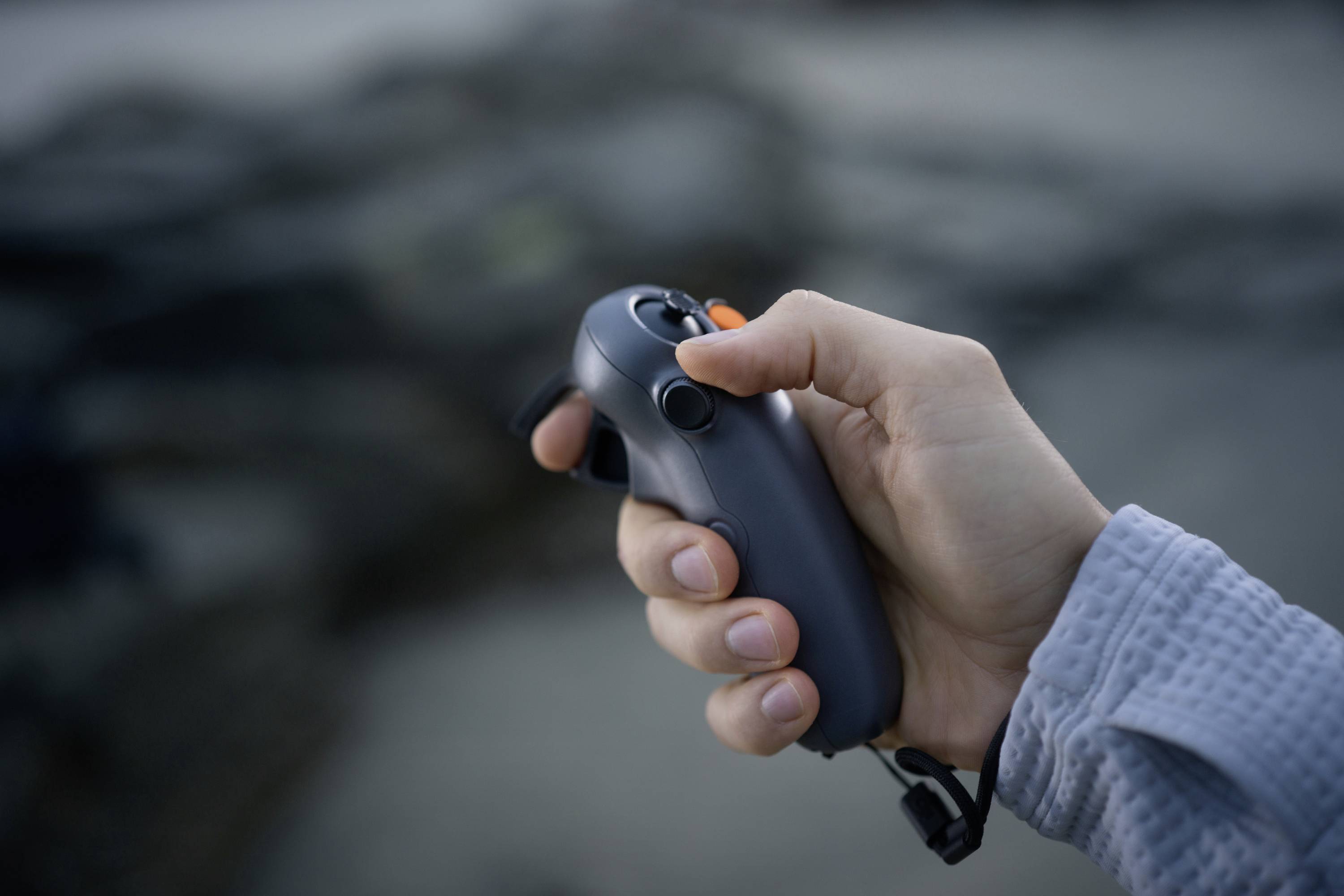 A hand is holding a remote control, with the thumb resting on a button. Background is out of focus.
