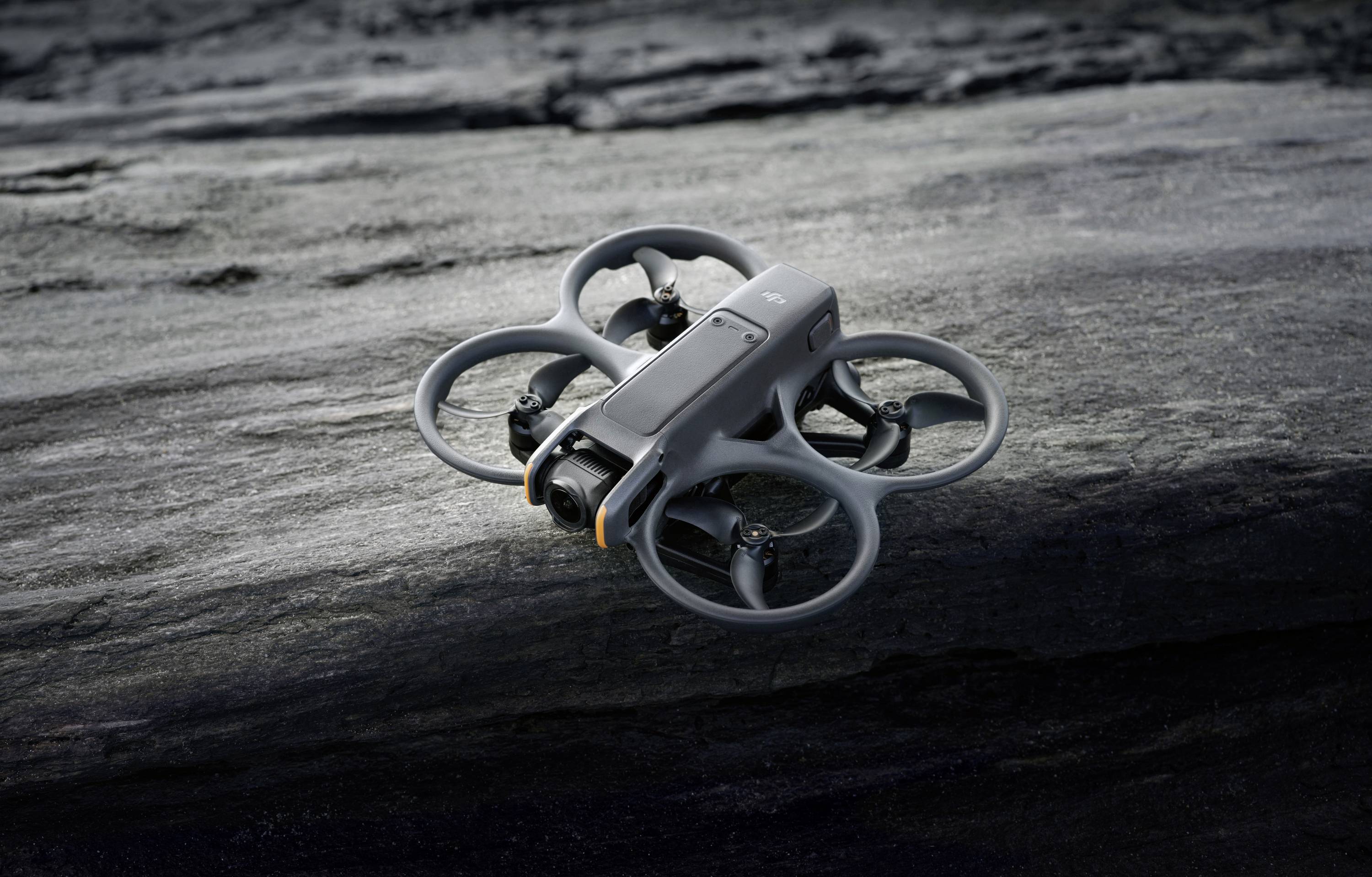 A grey drone rests on a rocky surface. It has four rotors with protective frames. The scene suggests an outdoor deployment.