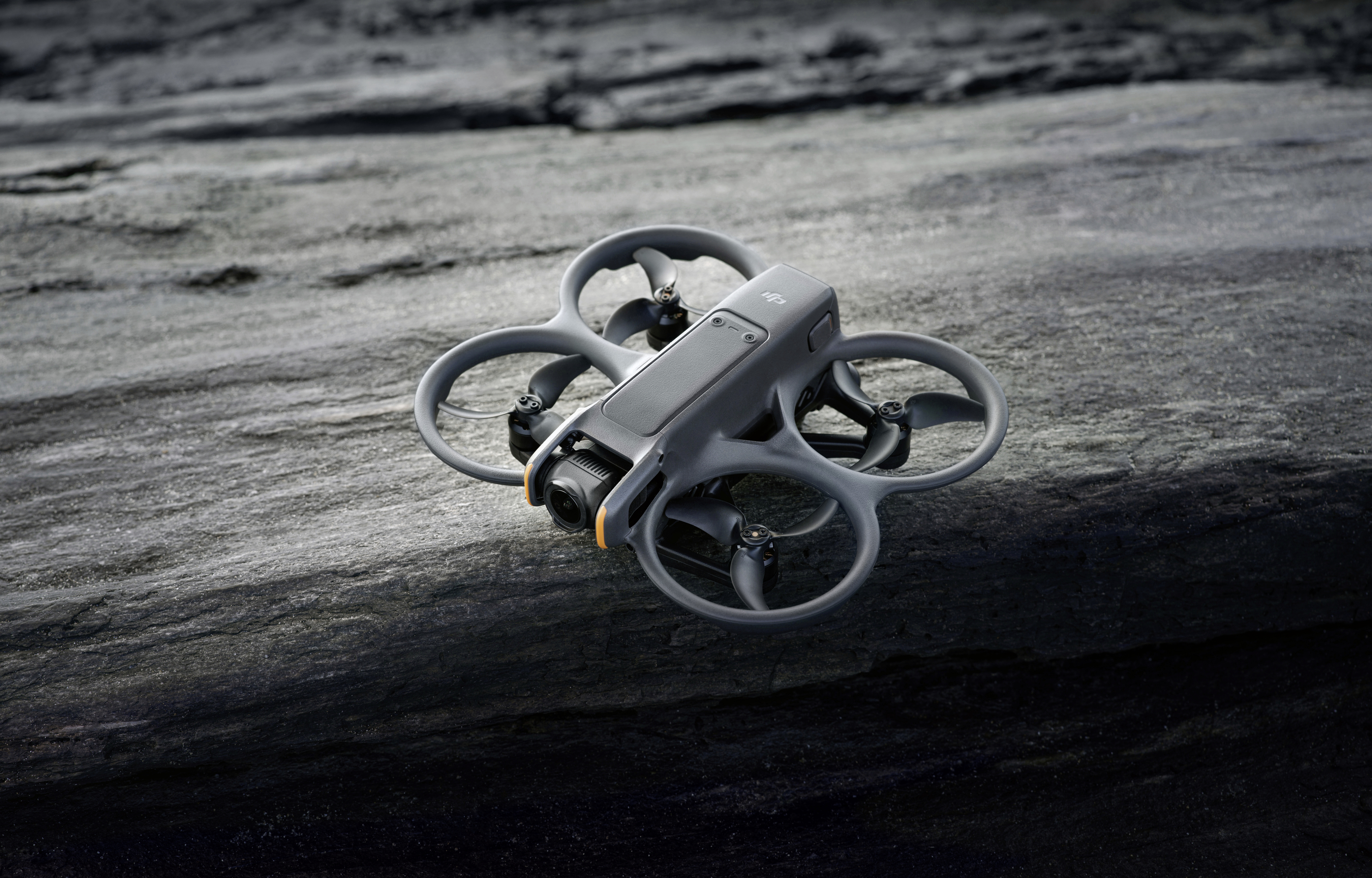 A grey drone rests on a rocky surface. It has four rotors with protective frames. The scene suggests an outdoor deployment.