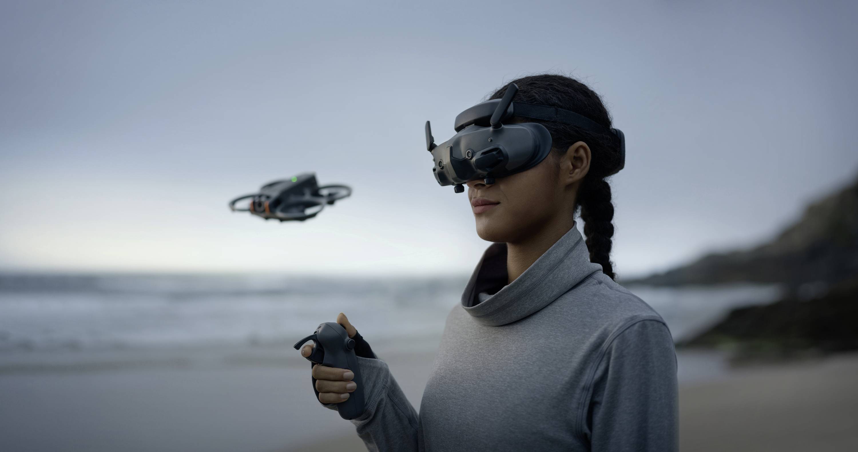 A woman is wearing a VR headset and controlling a small drone on the beach using a controller. The sky is cloudy.