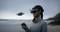 A woman is wearing a VR headset and controlling a small drone on the beach using a controller. The sky is cloudy.