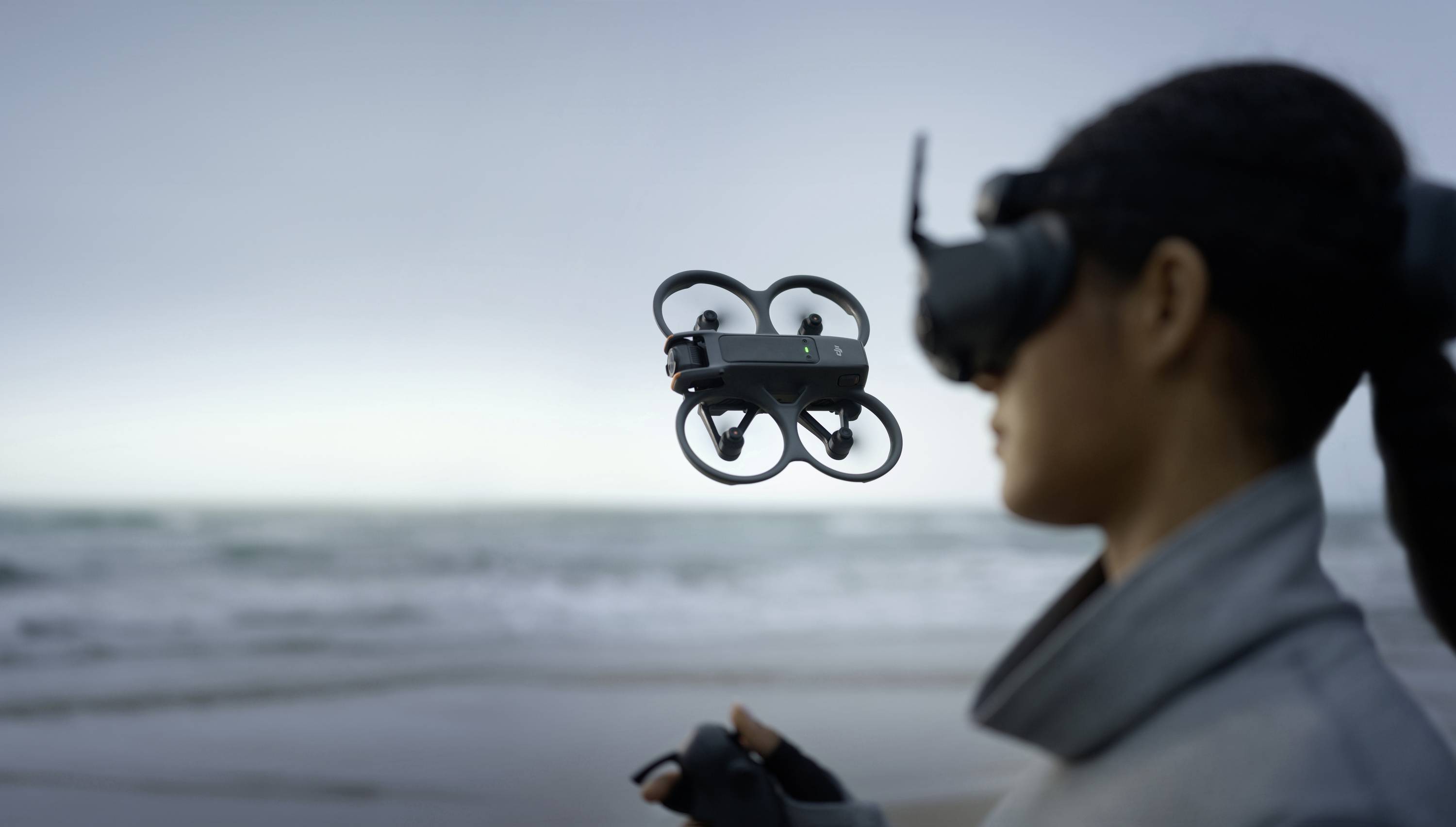 A person stands on the beach, controlling a small drone hovering in the air before them using a remote control and VR headset.