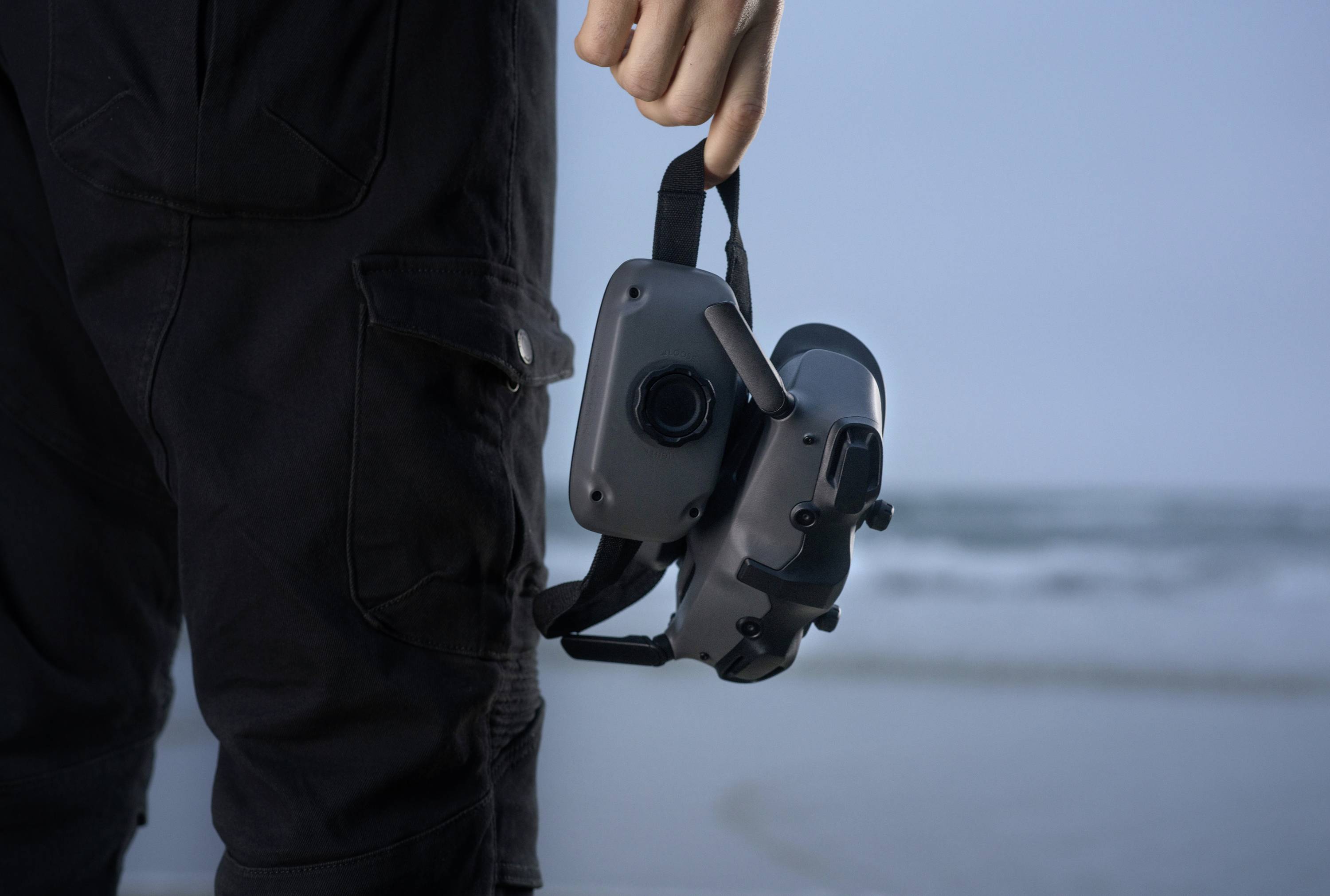 A person is holding a VR headset while standing on a beach. Blurred seascape is visible in the background.