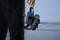 A person is holding a VR headset while standing on a beach. Blurred seascape is visible in the background.