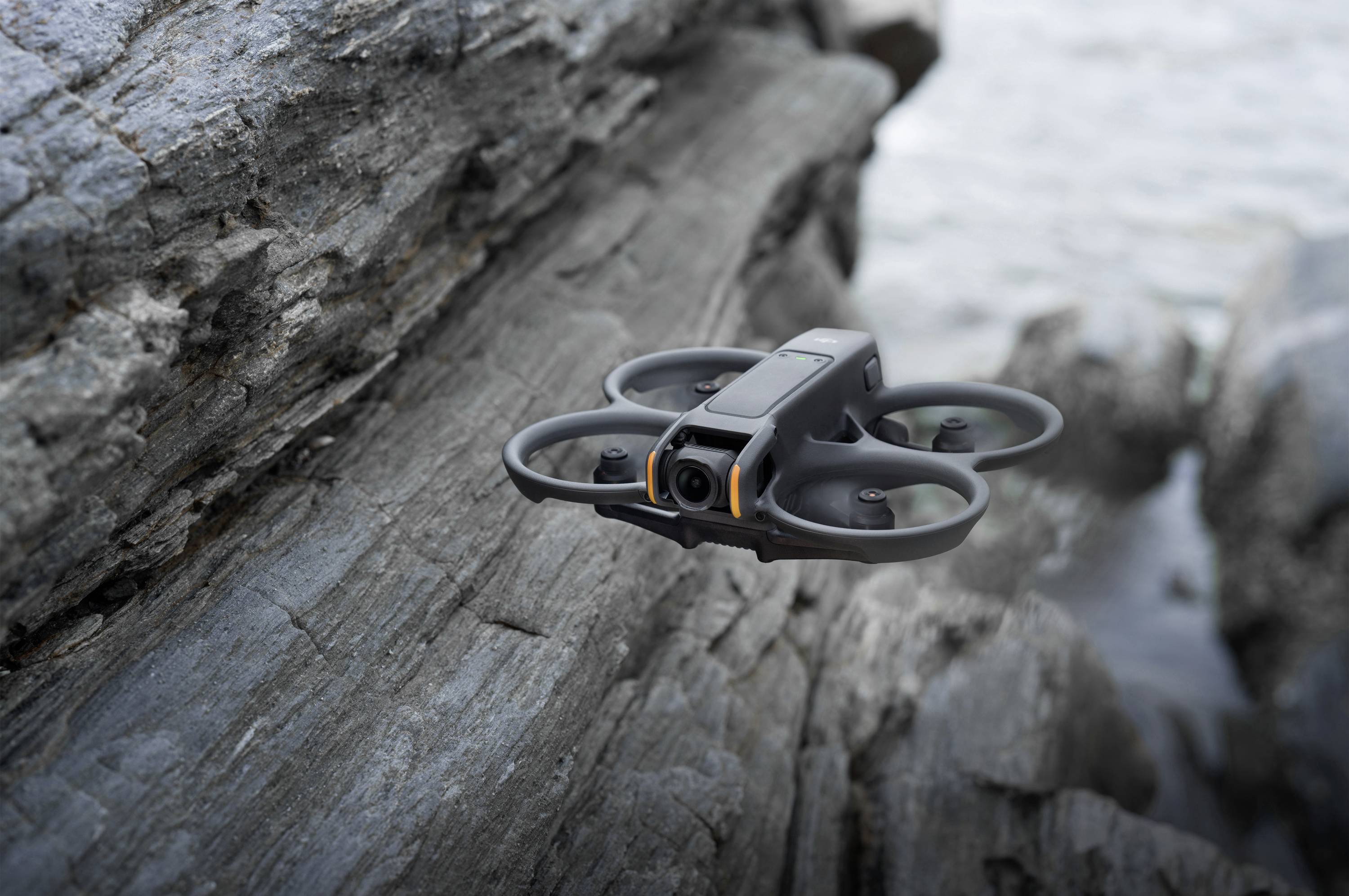 A black drone flies close to a rocky cliff. The sea is blurrily visible in the background.