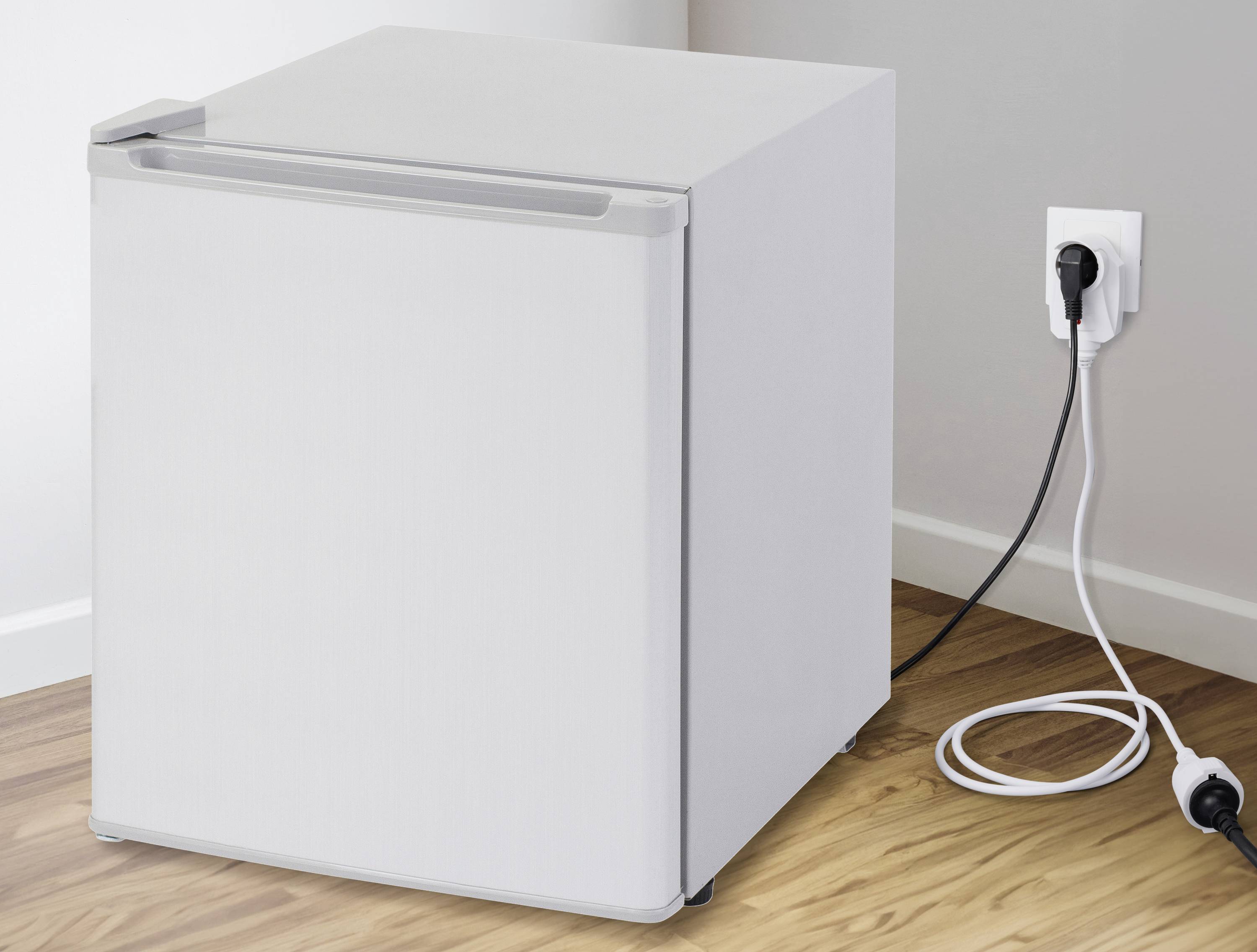 A white mini-fridge is standing on a wooden floor and is plugged into a power socket.
