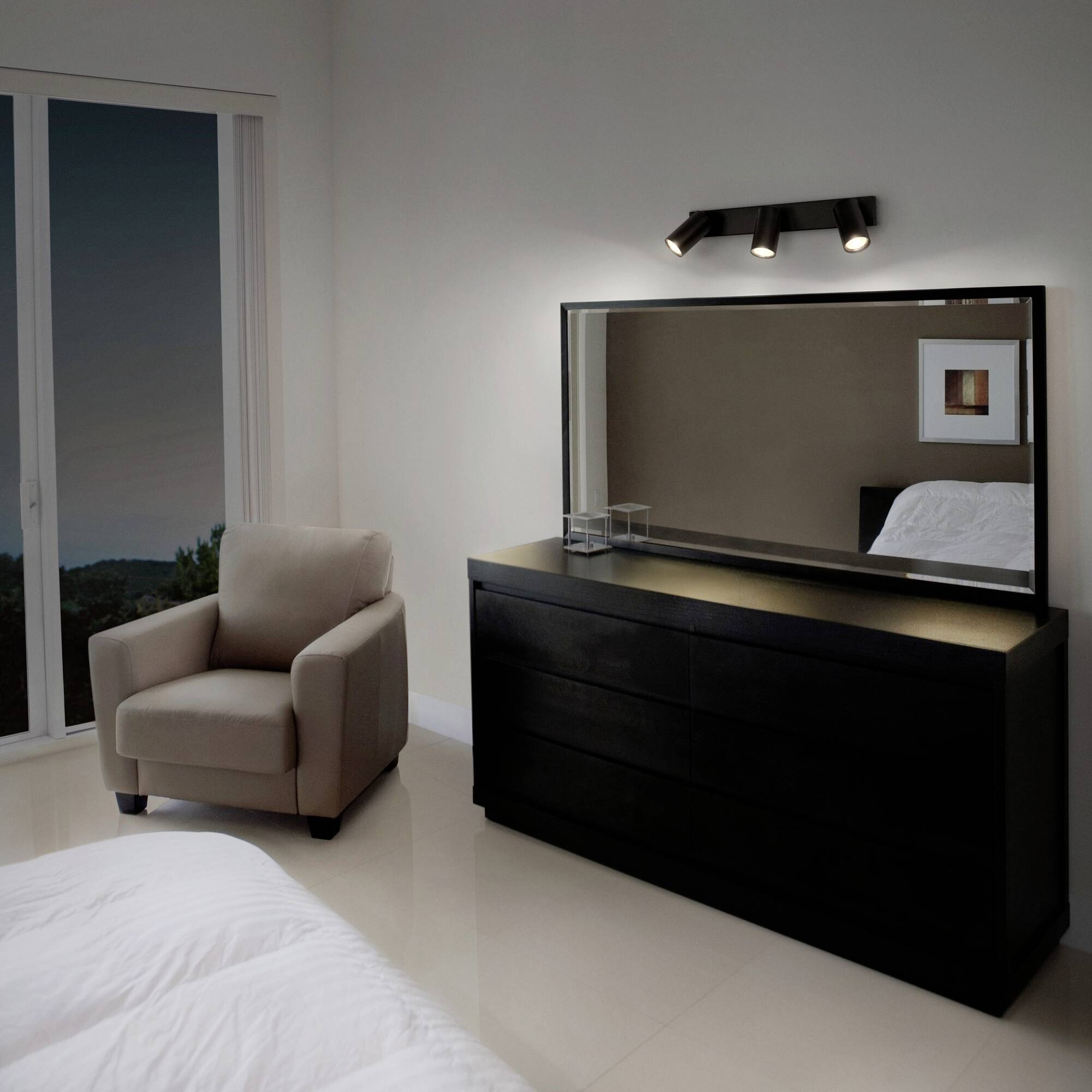A modern bedroom with a large mirror, an armchair, and a window offering a nocturnal view.