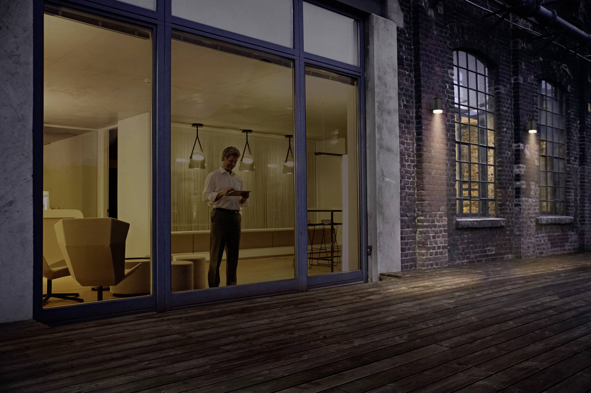 A person stands in the evening in a modern, well-lit room behind large glass windows. Outside it is dark, with an illuminated wooden deck.