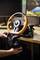A person is attaching an orange and black steering wheel to a desk. A monitor and keyboard are visible in the background.