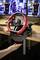 A person is installing a red and black racing steering wheel at a desk, with two monitors and gaming chairs in the background.