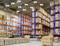 A large warehouse with tall shelving, filled with boxes and parcels. Bright industrial lighting and pallets on the floor.