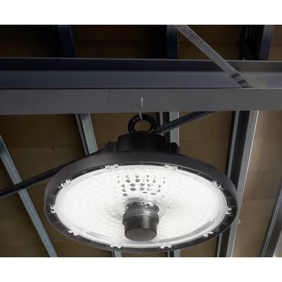 A round LED lamp hangs from a metal support ceiling. The lamp is switched on and illuminating an industrial environment.