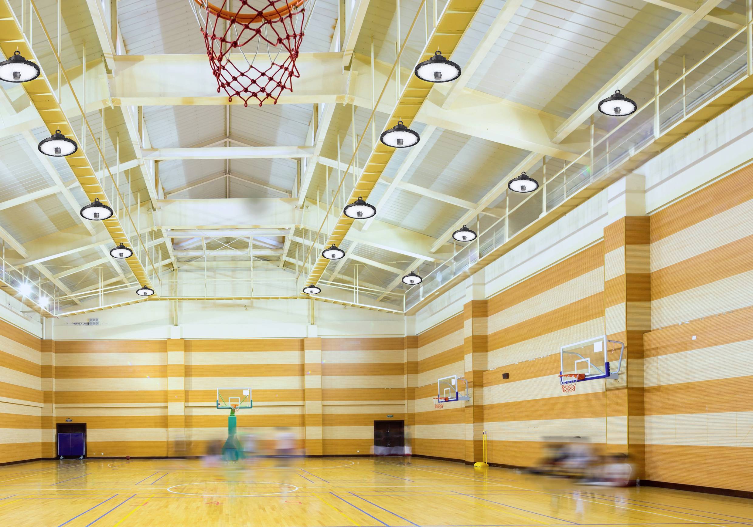 An empty sports hall with a wooden floor and basketball hoops on the sides. The ceiling is high with exposed beams and lights.