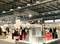 Exhibition hall with visitors examining stands displaying furniture and home accessories. Bright lighting and modern displays.